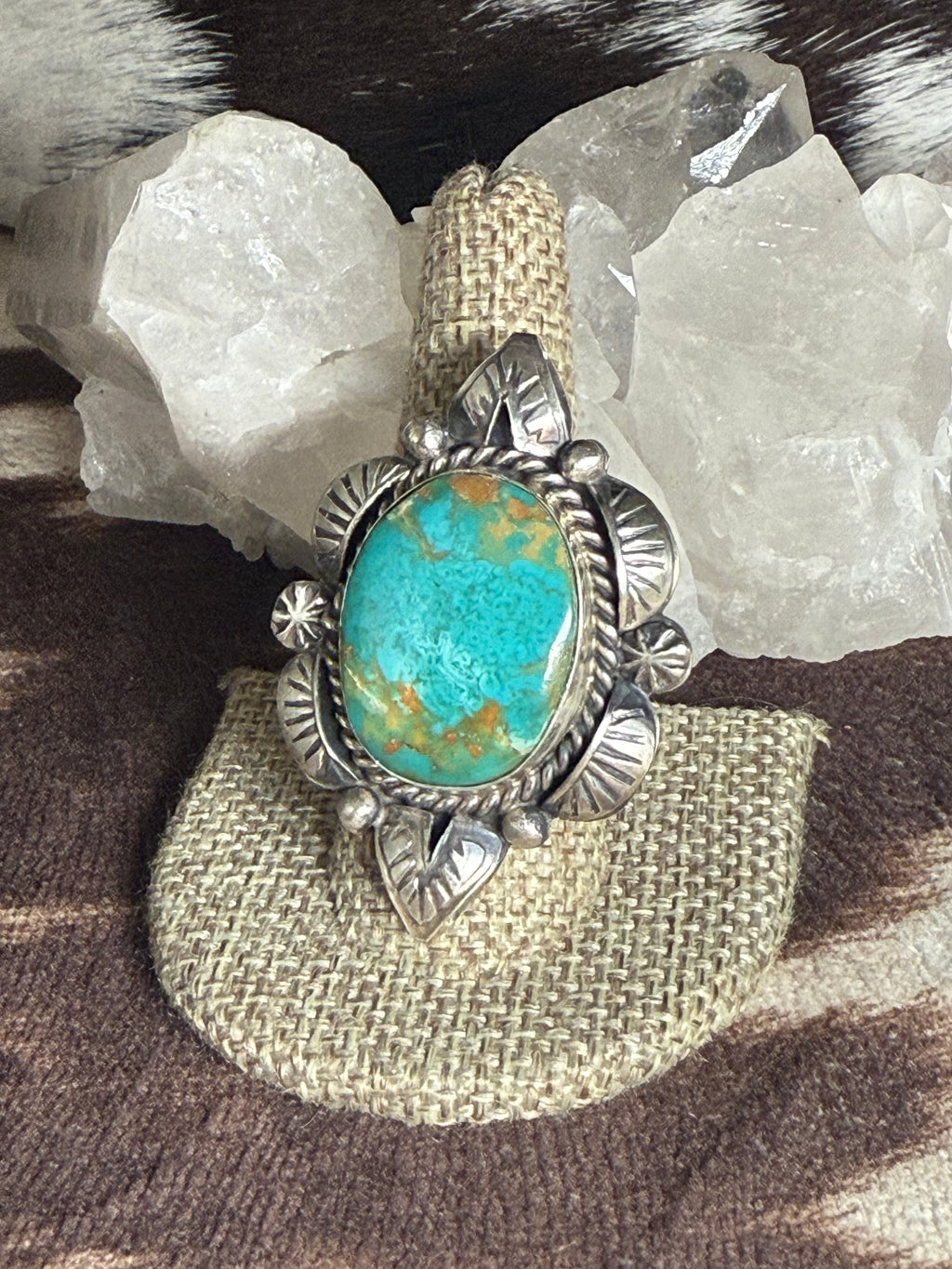 A sterling silver ring featuring a large oval turquoise cabochon with natural brown matrix, displayed on a woven beige ring stand with white quartz crystals and a brown-and-white cowhide surface in the background.