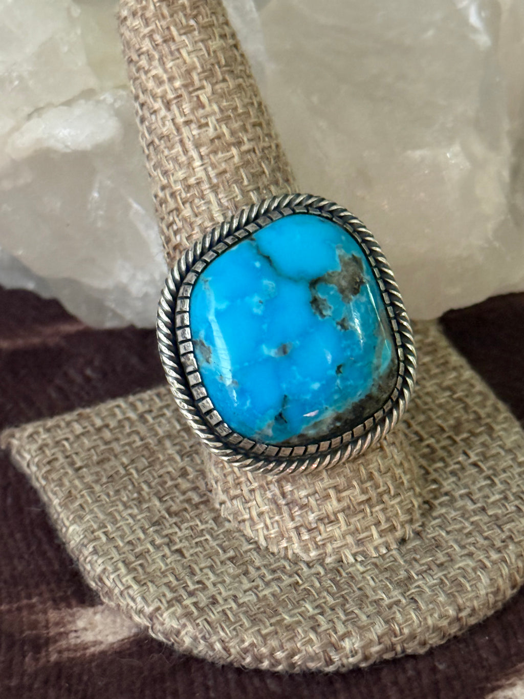 A silver ring with a large oval turquoise stone, displayed on a woven ring stand with a white crystal in the background.