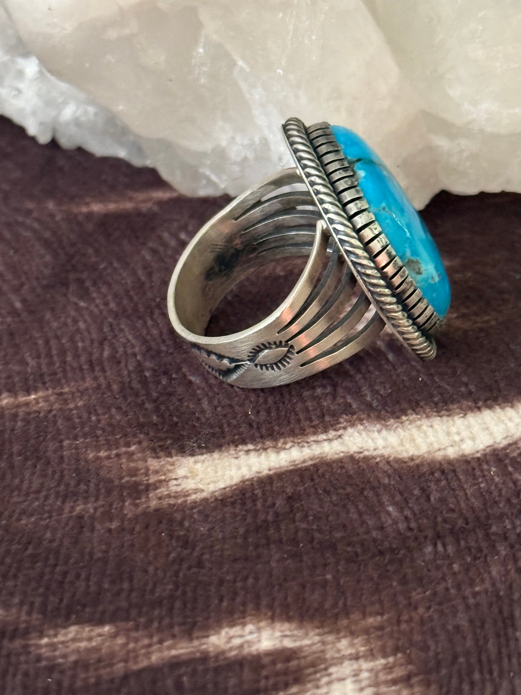 A silver-toned ring with a large, vibrant turquoise blue oval stone, displayed on a textured brown surface with a white crystal in the background.