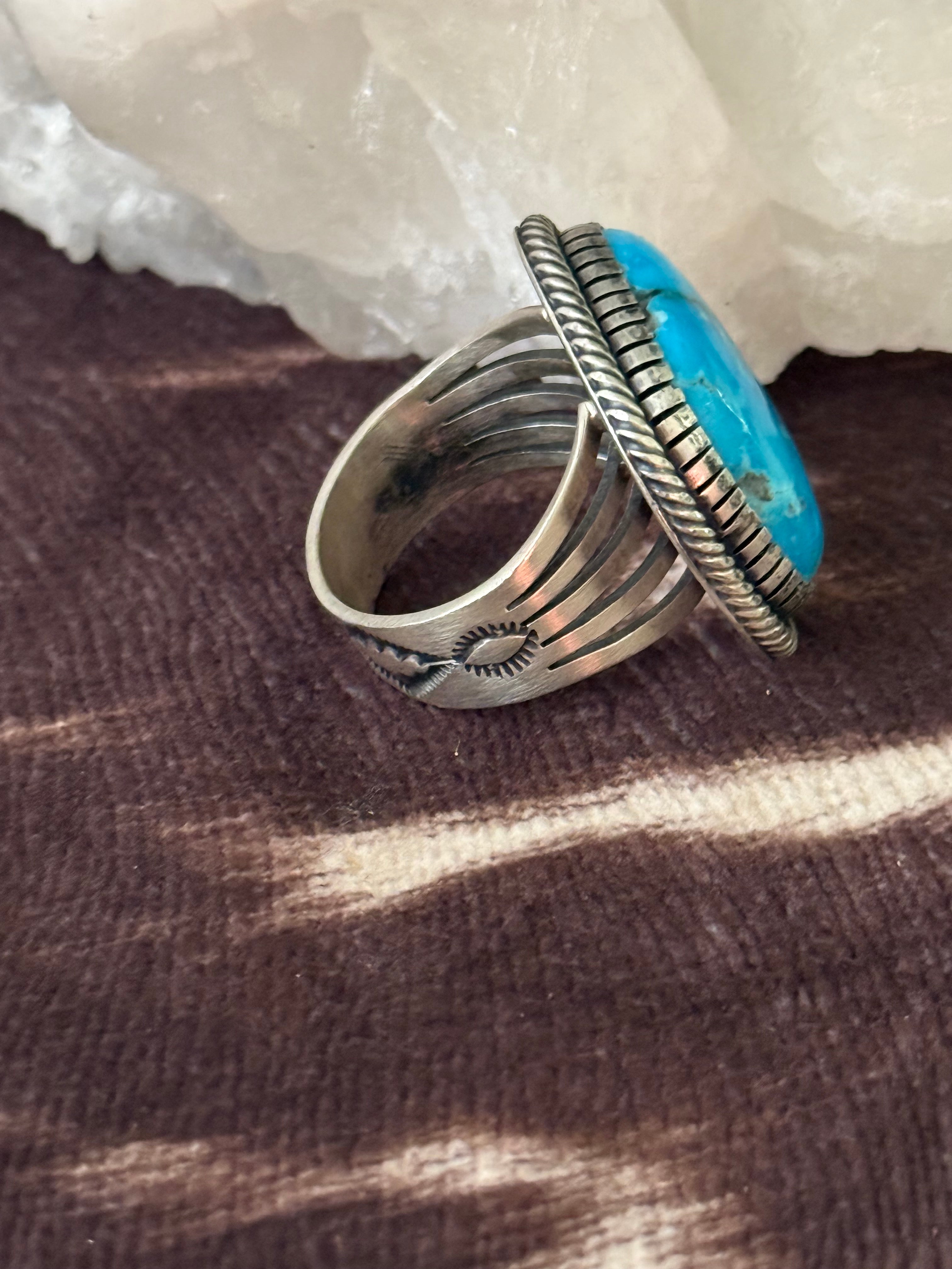 A silver-toned ring with a large, vibrant turquoise blue oval stone, displayed on a textured brown surface with a white crystal in the background.