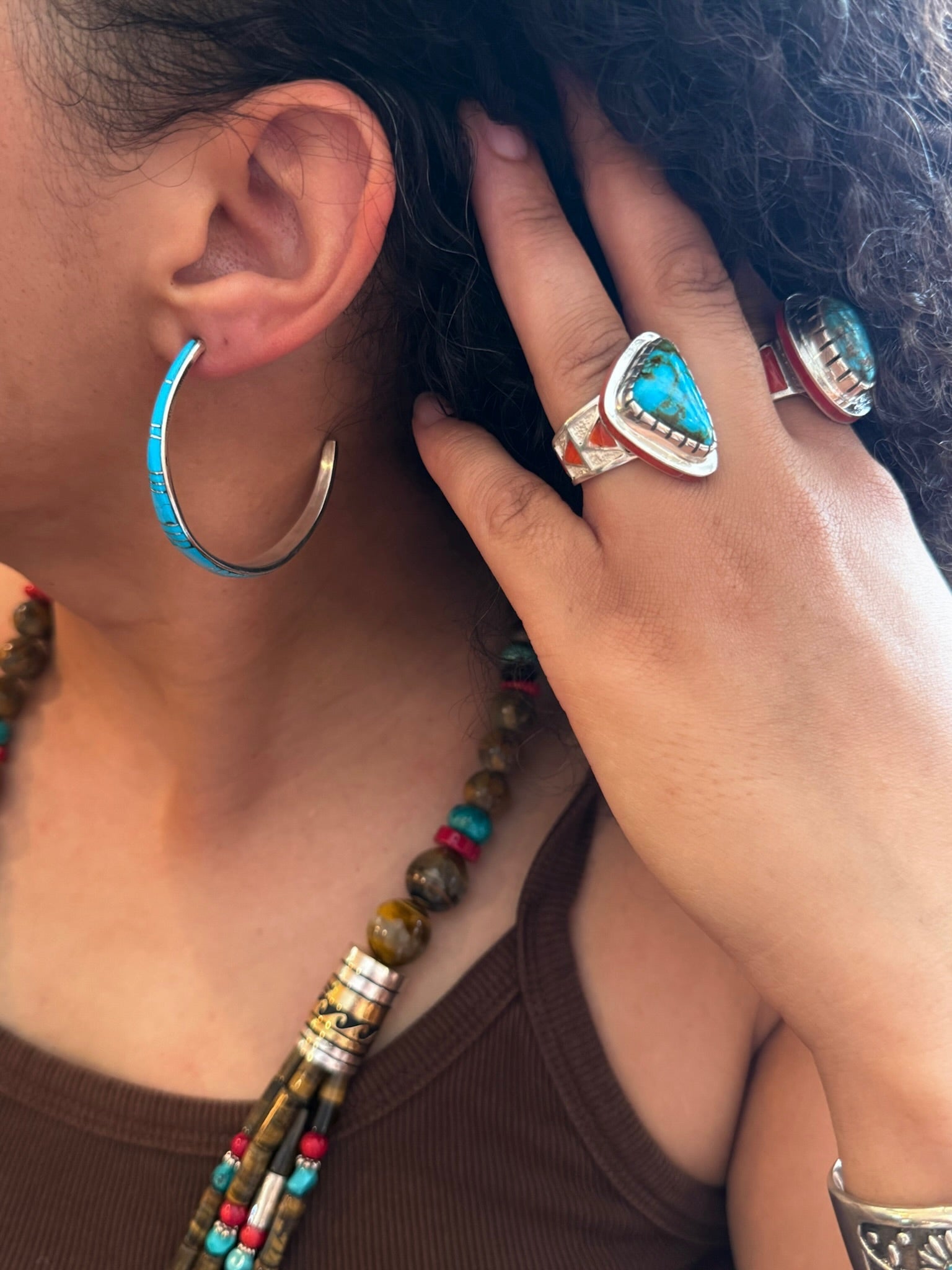 A close-up lifestyle shot of a person wearing multiple turquoise and silver jewelry pieces, including a large silver hoop earring with turquoise inlay, a multi-strand beaded necklace, and two silver rings with large turquoise stones.