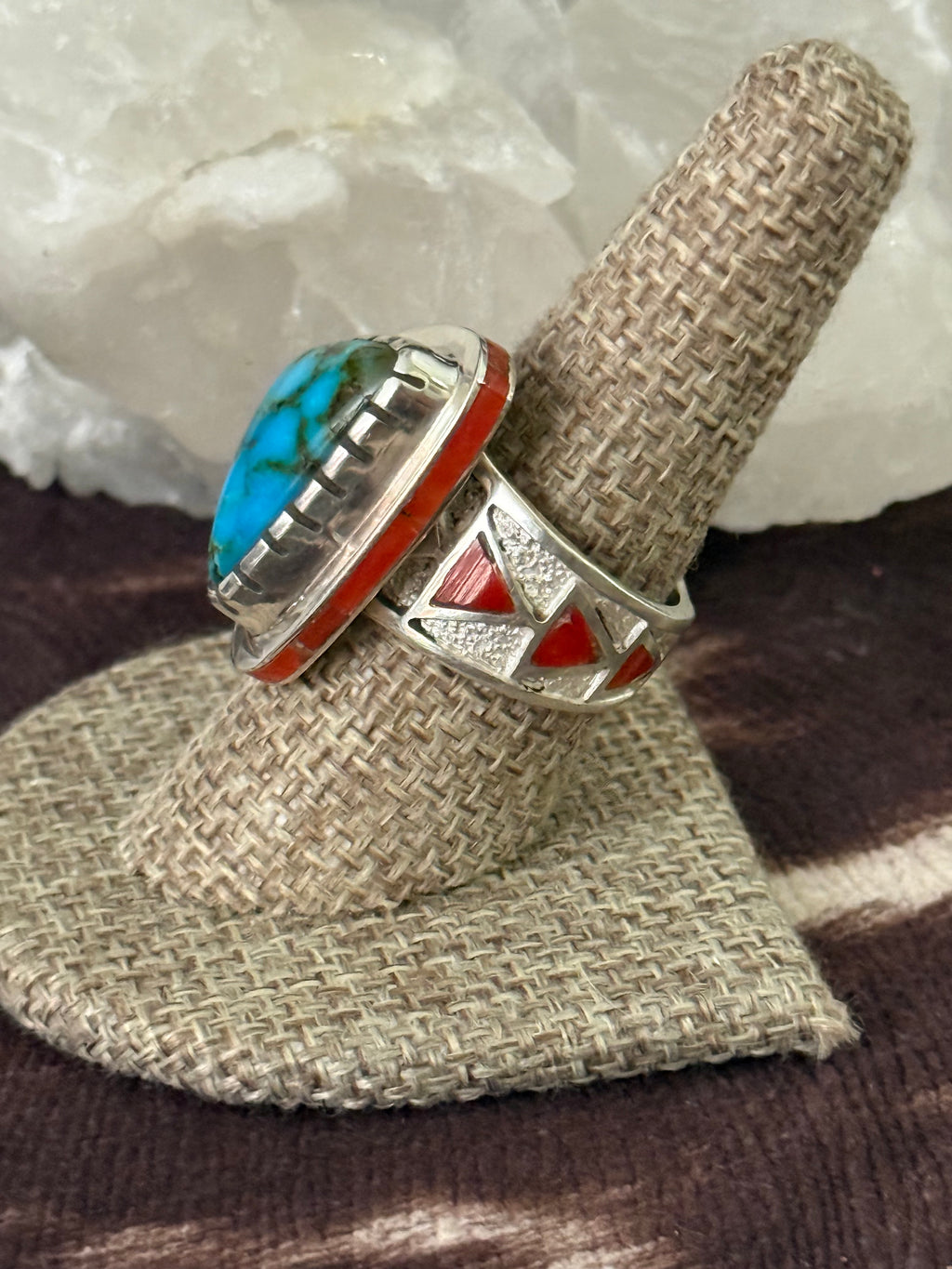 A sterling silver ring with a turquoise and red coral inlay, displayed on a woven ring stand.