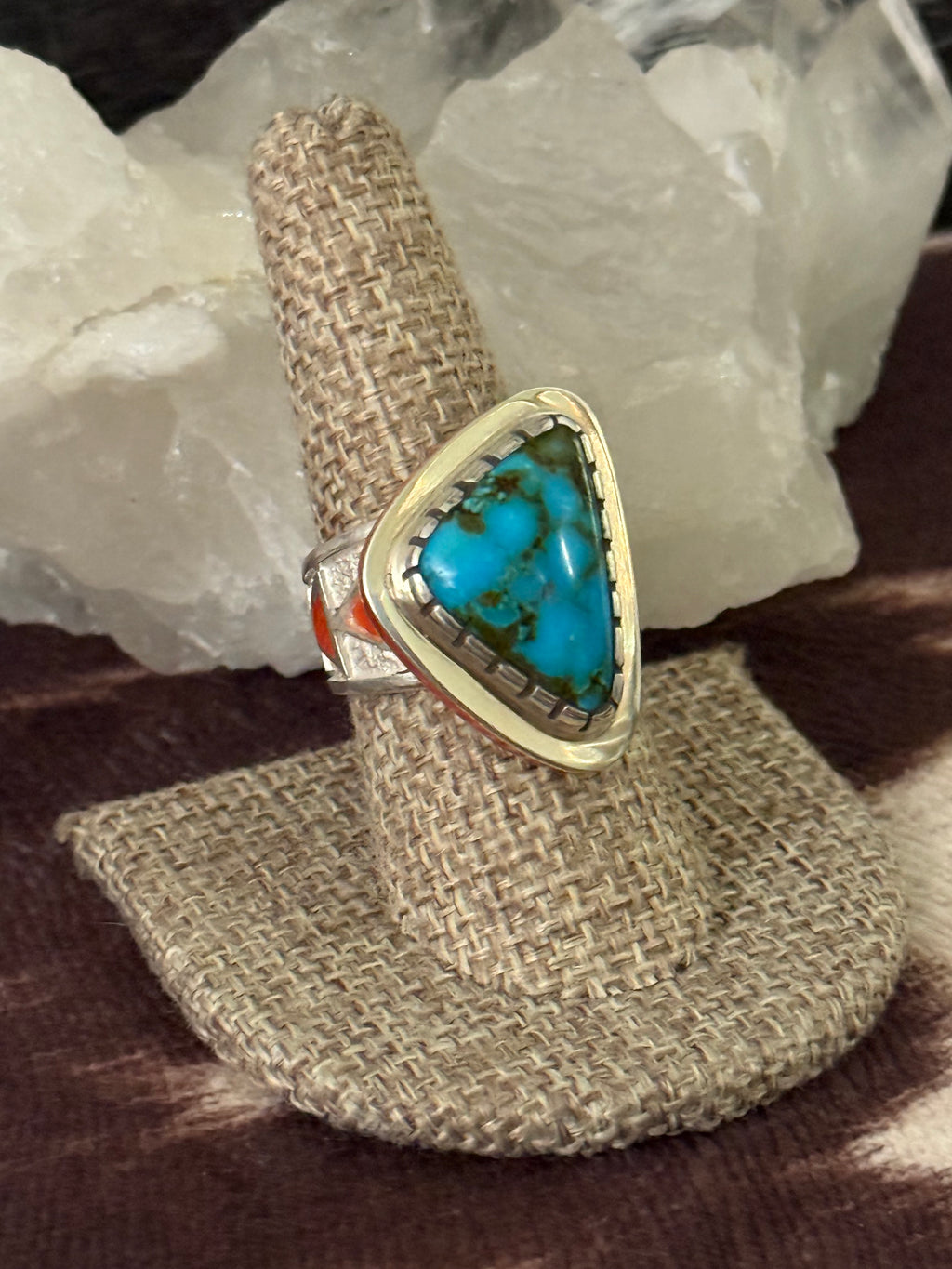 A triangular turquoise ring with a gold-toned bezel and silver-toned band details, displayed on a woven ring stand with white crystal rocks in the background.