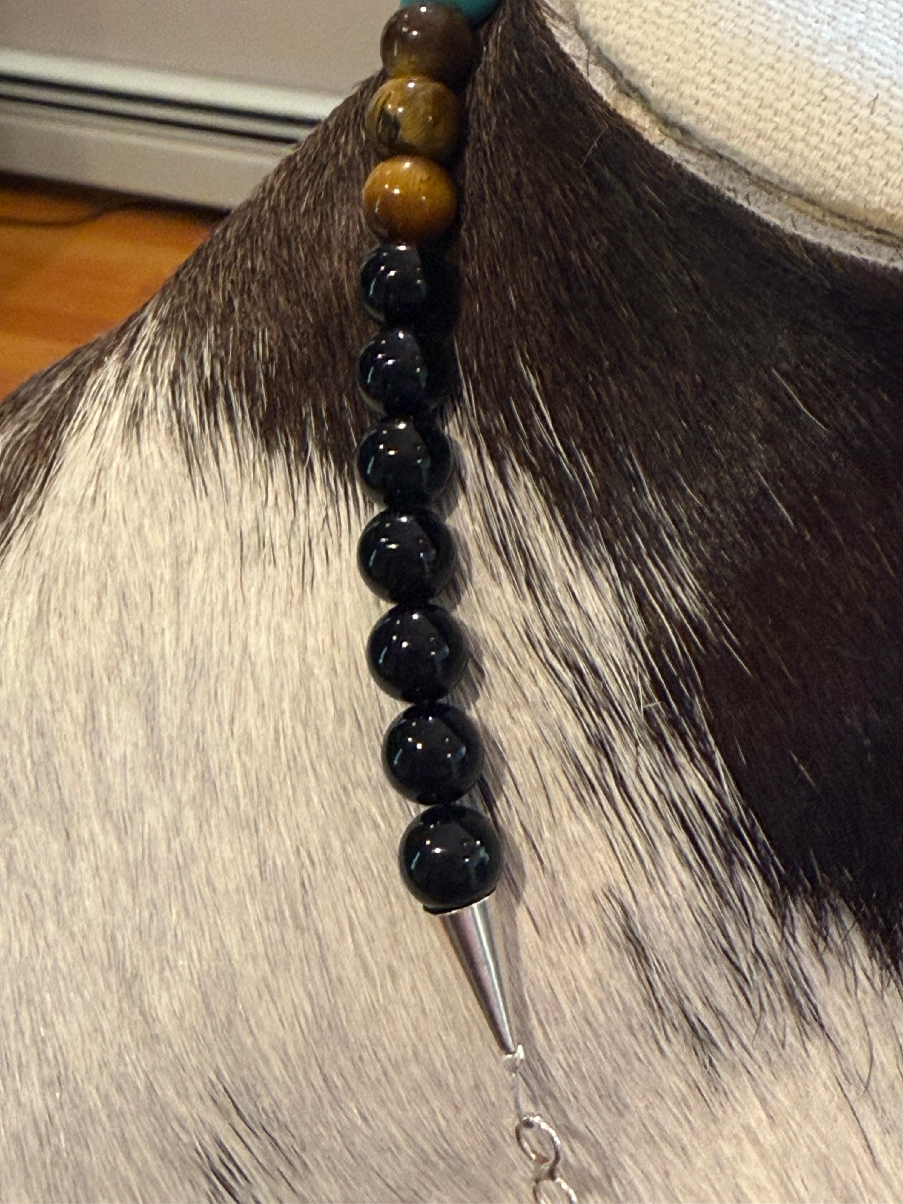 A close-up of a beaded necklace draped over a cowhide mannequin. The necklace features a vertical strand of glossy black beads, a single round brown tiger's eye bead, and a turquoise accent bead at the top, finished with a silver conical spike clasp.