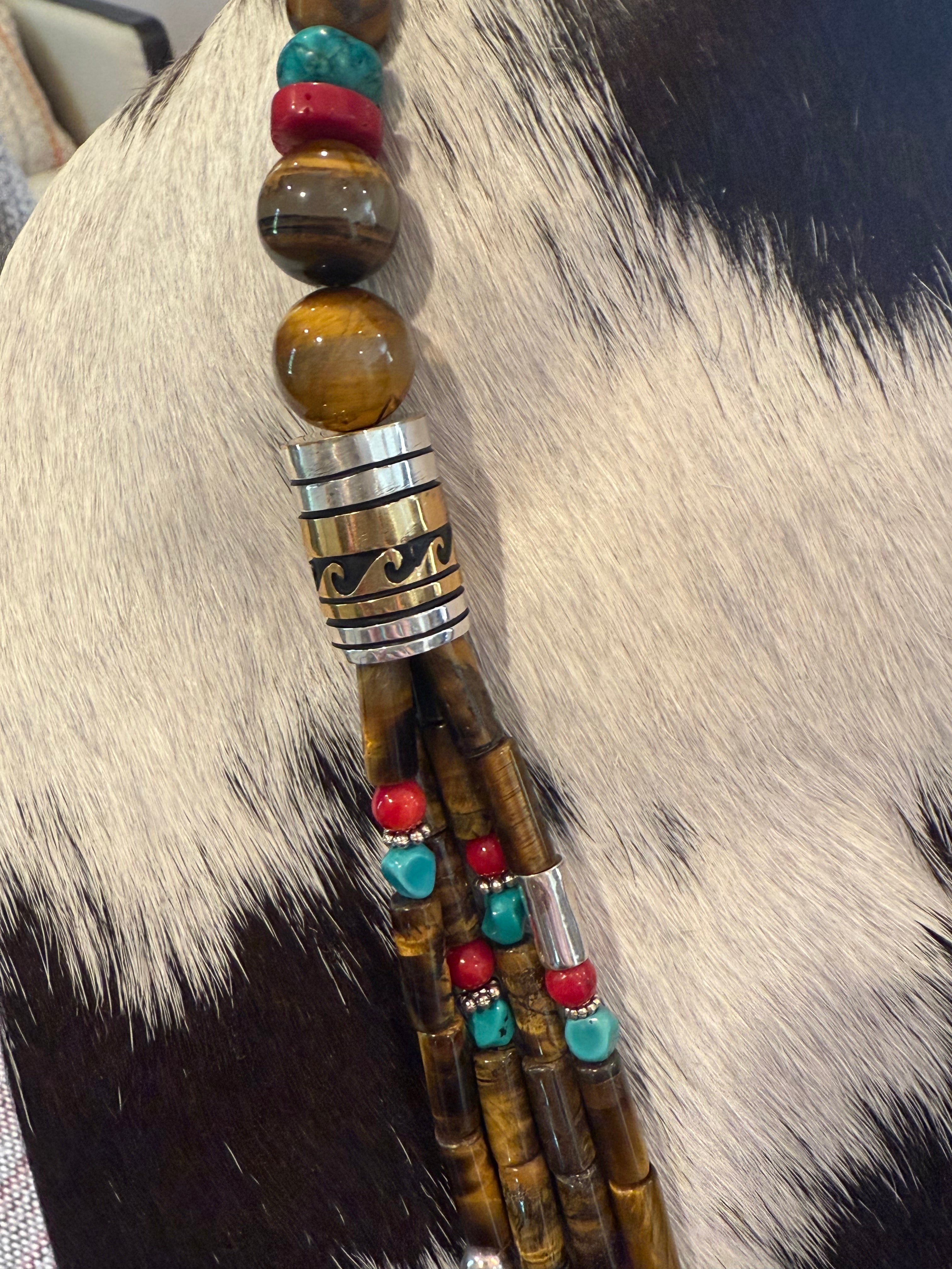 A close-up of a beaded necklace draped over cowhide. The necklace features large polished tiger's eye beads, a silver-toned cylindrical spacer with black engraved patterns, and dangling strands of brown beads accented with turquoise and red beads.