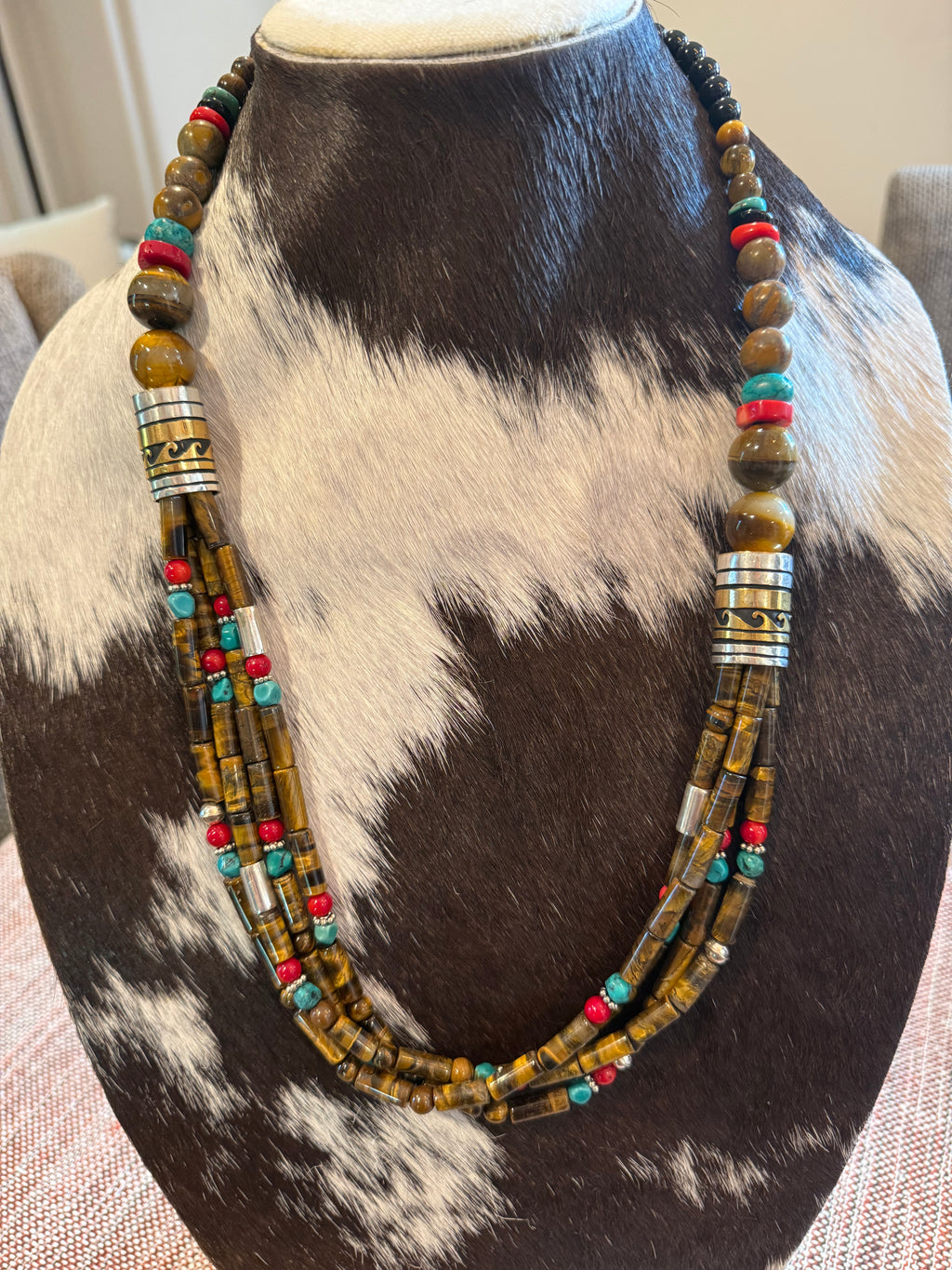A multi-strand beaded necklace displayed on a cowhide mannequin bust. The necklace features brown and gold tiger's eye beads, silver-toned cylindrical spacer beads, and small turquoise and red accent beads.