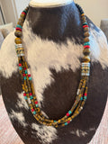 A multi-strand beaded necklace displayed on a cowhide mannequin bust. The necklace features brown and gold tiger's eye beads, silver-toned cylindrical spacer beads, and small turquoise and red accent beads.