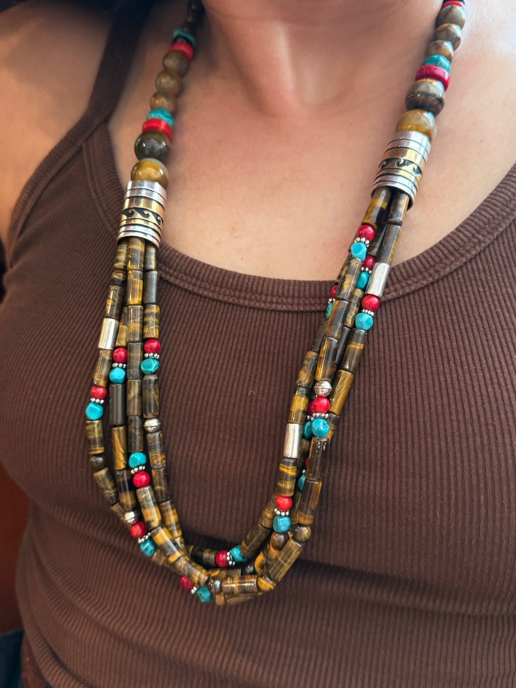 A close-up of a person wearing a multi-strand statement necklace made of tiger's eye beads, metallic silver spacers, and small turquoise and red beads, paired with a brown ribbed tank top.
