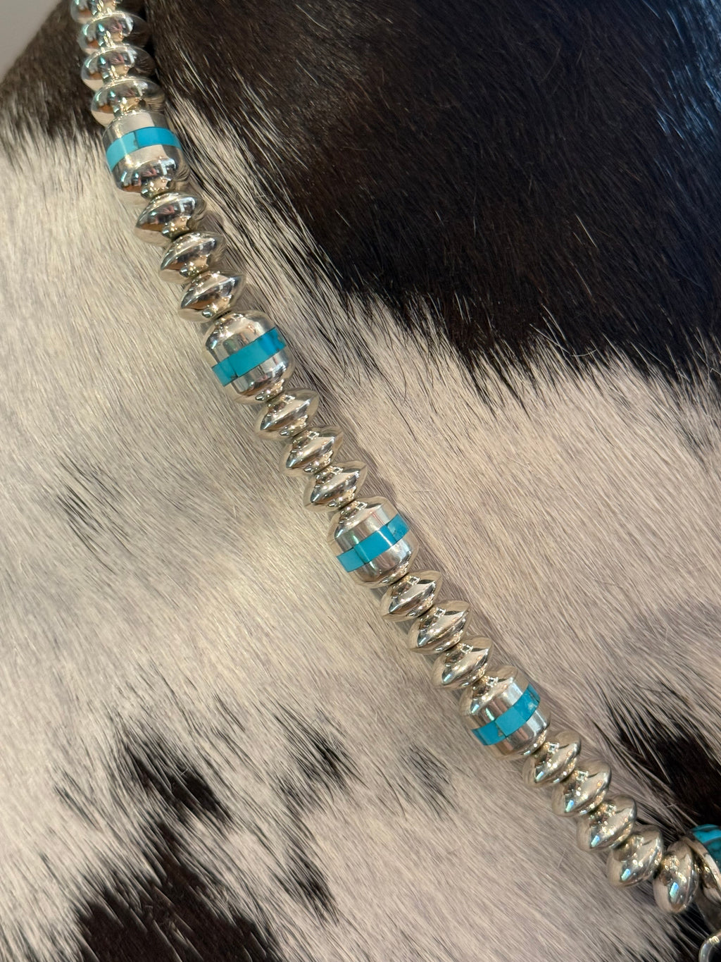 A close-up of a beaded necklace featuring silver-toned disc-like beads and bright turquoise blue spacer bands, laid diagonally across a black-and-white cowhide surface.
