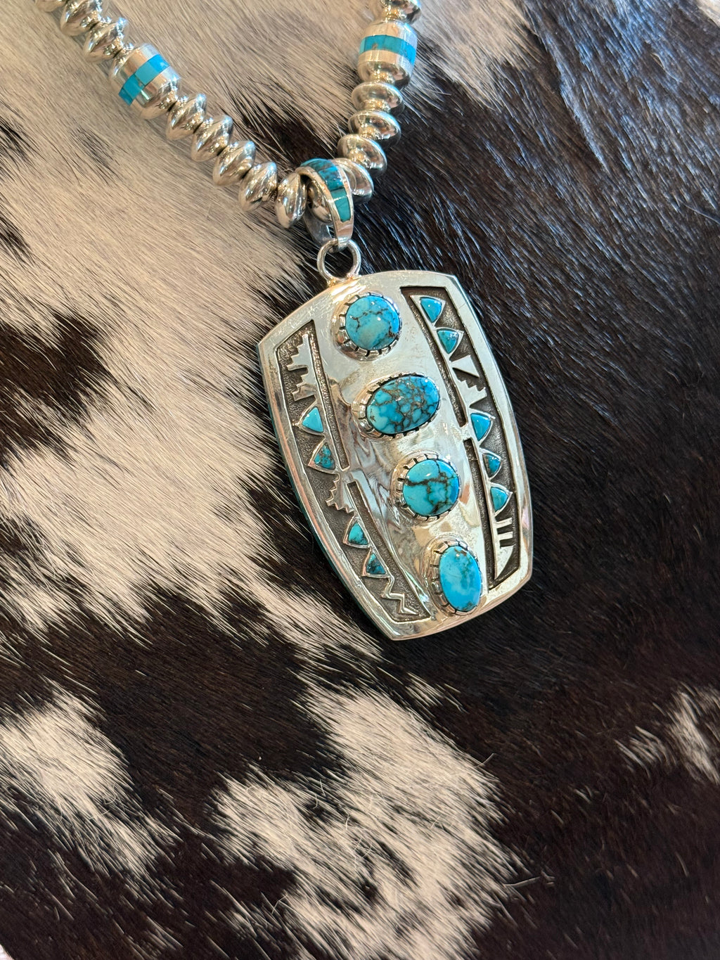 A silver pendant necklace with turquoise stones, displayed on a black and white cowhide background.