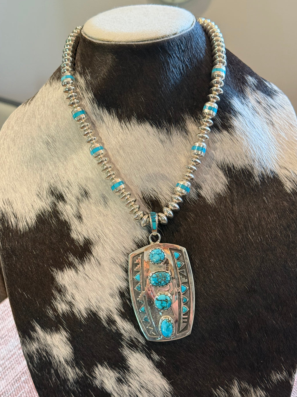 A silver-toned beaded necklace with turquoise blue stones, displayed on a cowhide mannequin bust. The necklace features a large rectangular pendant with four vertical turquoise stones and silver geometric detailing.