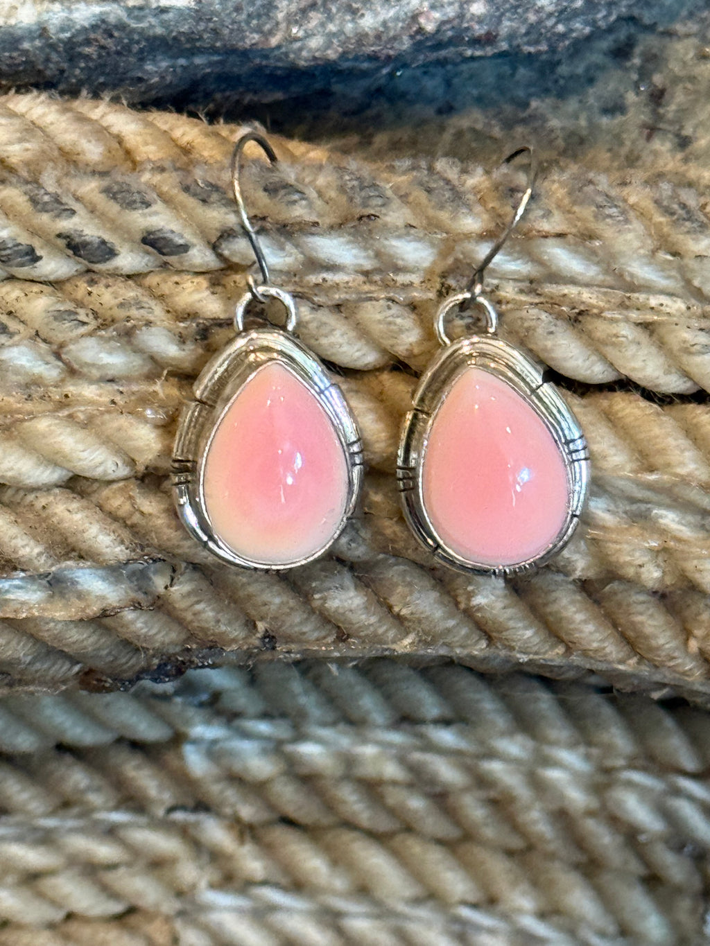 A pair of silver drop earrings with teardrop-shaped pink cabochon stones, displayed on a textured beige rope background.