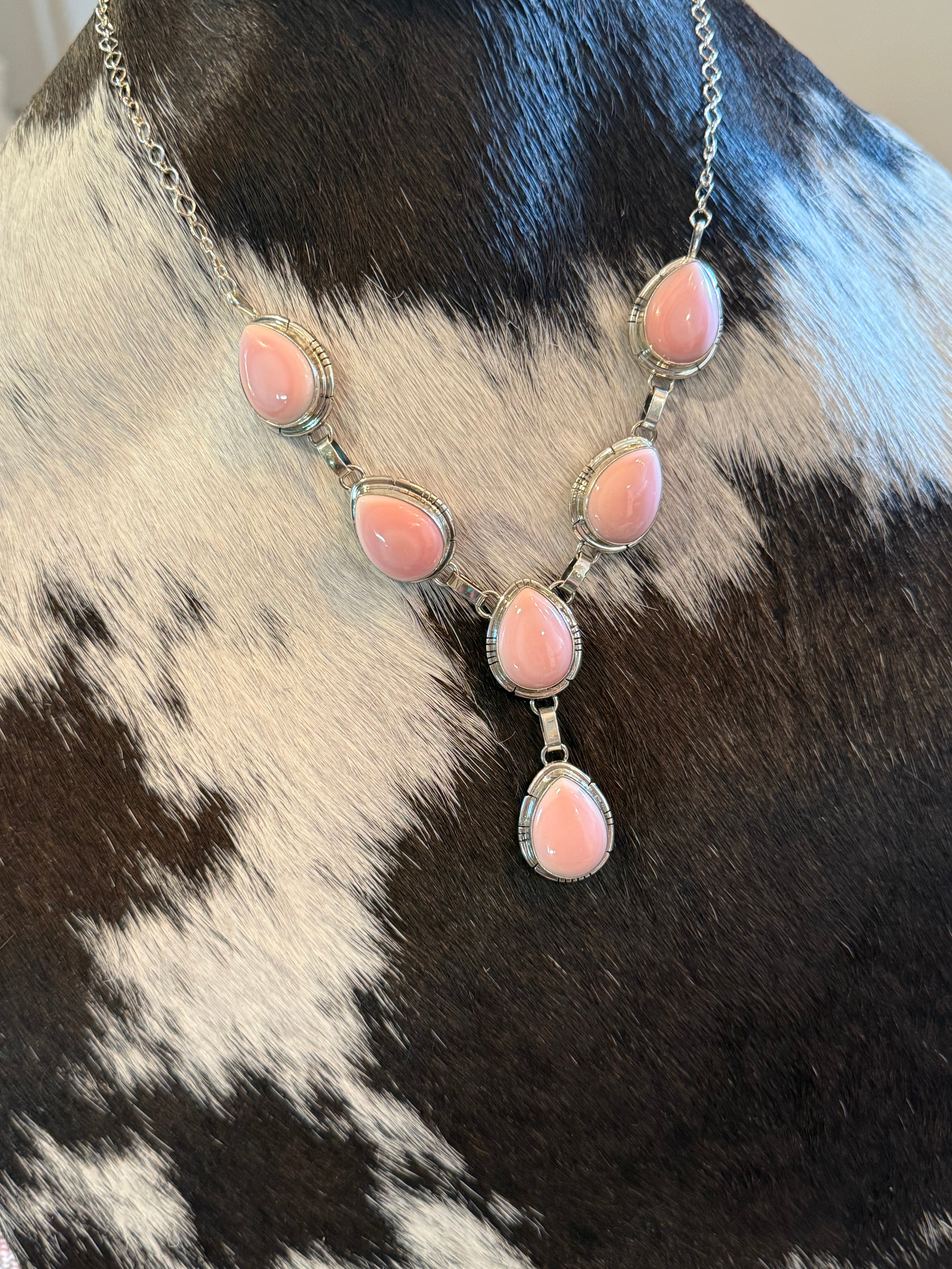 A silver-toned statement necklace with five pink teardrop-shaped stones, displayed on a black and white cowhide surface.