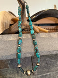 A beaded necklace with turquoise-blue stone beads and small metallic bronze-toned spacer beads, draped over a rustic wooden stand in front of a stone surface.