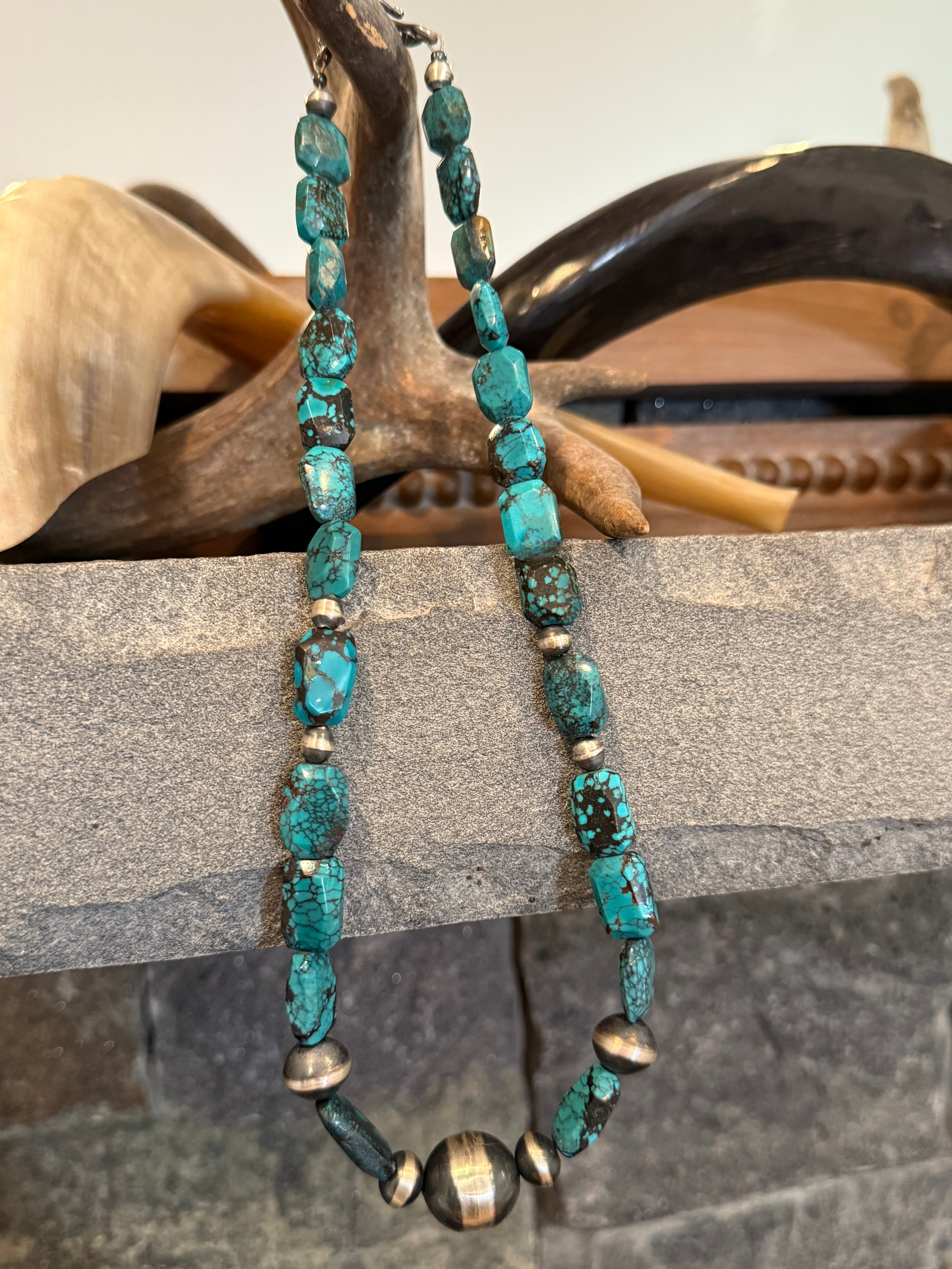 A beaded necklace with turquoise-blue stone beads and small metallic bronze-toned spacer beads, draped over a rustic wooden stand in front of a stone surface.