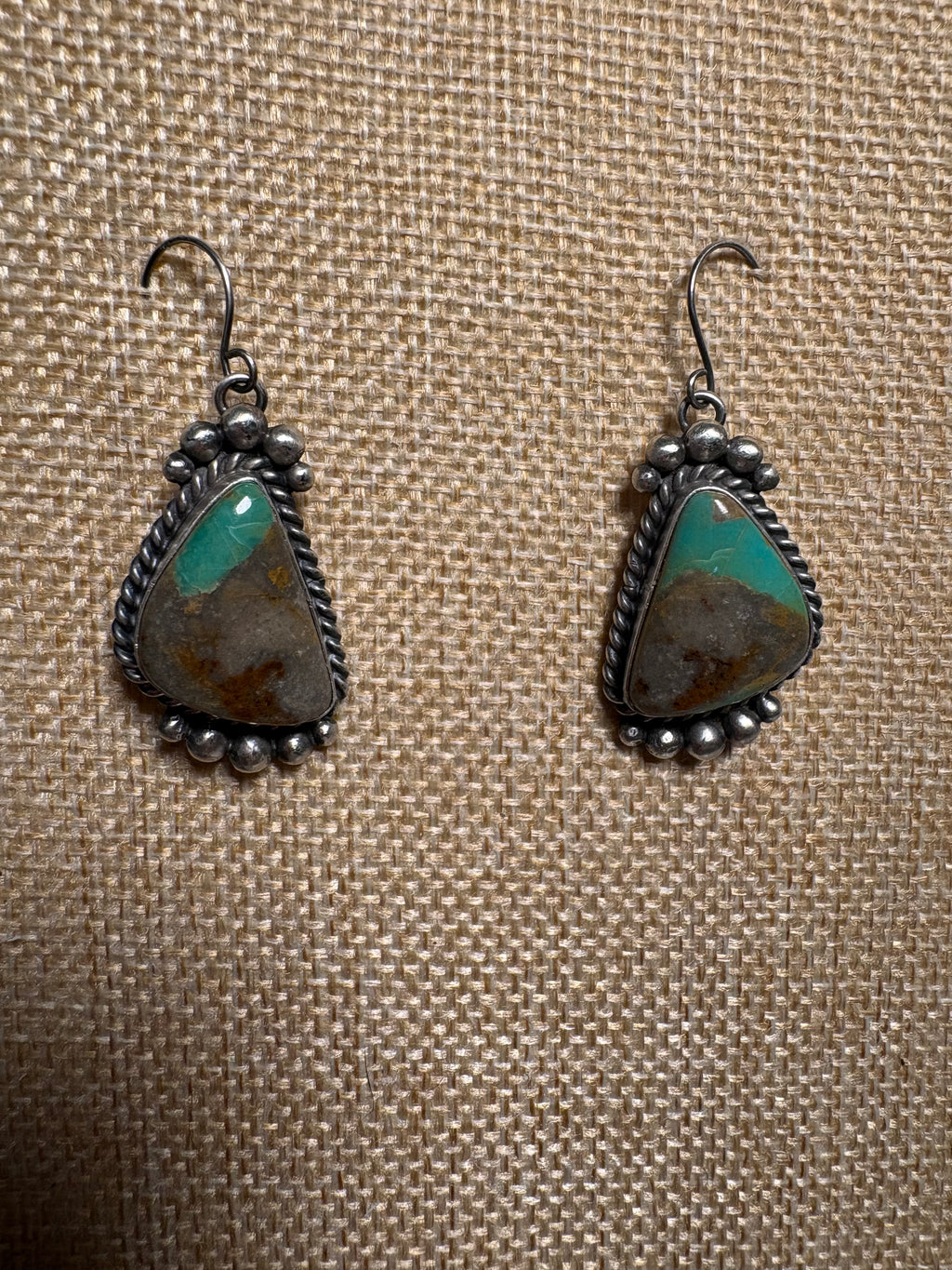 A pair of handmade silver dangle earrings with turquoise and brown stone inlays, displayed on a textured beige burlap background.