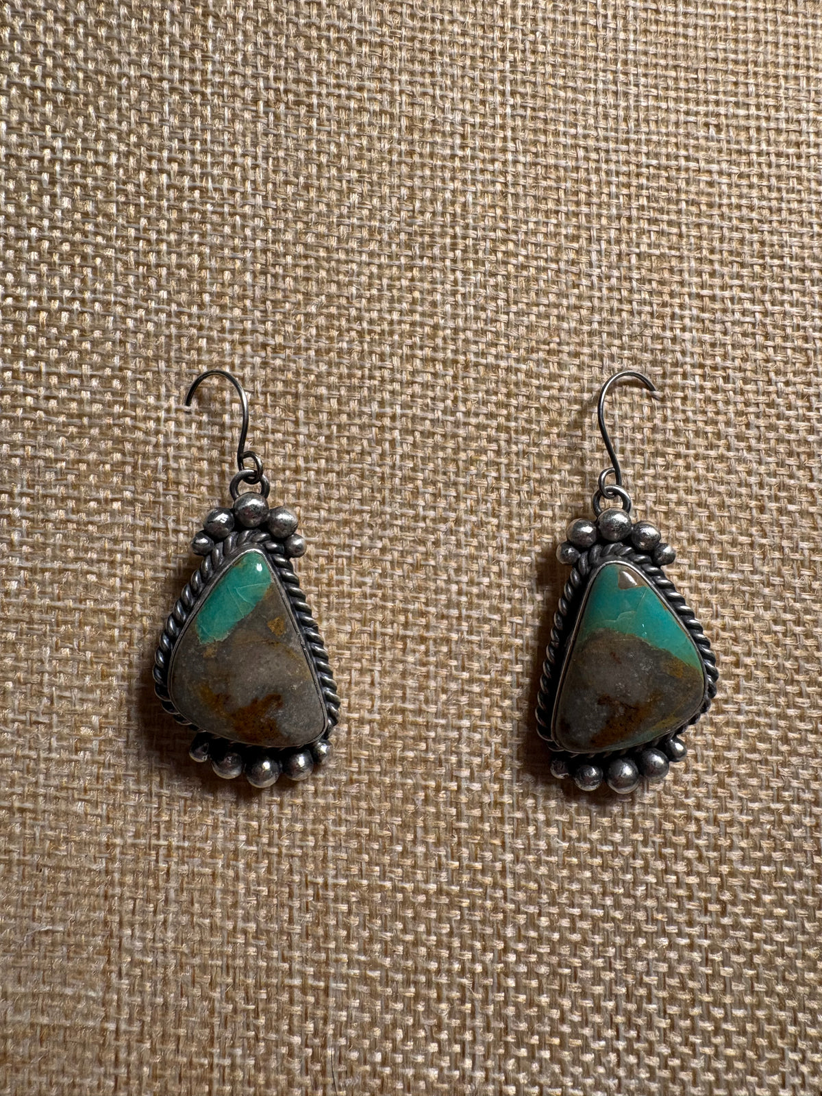 A pair of handmade sterling silver dangle earrings with teardrop-shaped turquoise and brown stone inlays, displayed on a textured beige burlap background.