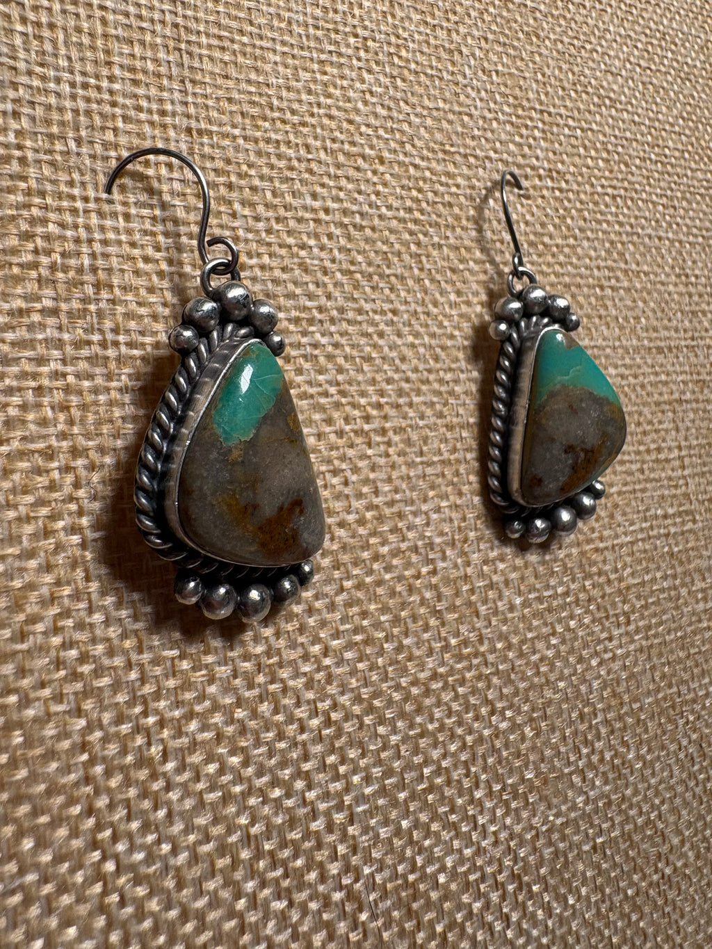 A pair of handmade sterling silver dangle earrings featuring natural turquoise and brown stone inlays, displayed on a textured beige burlap background.