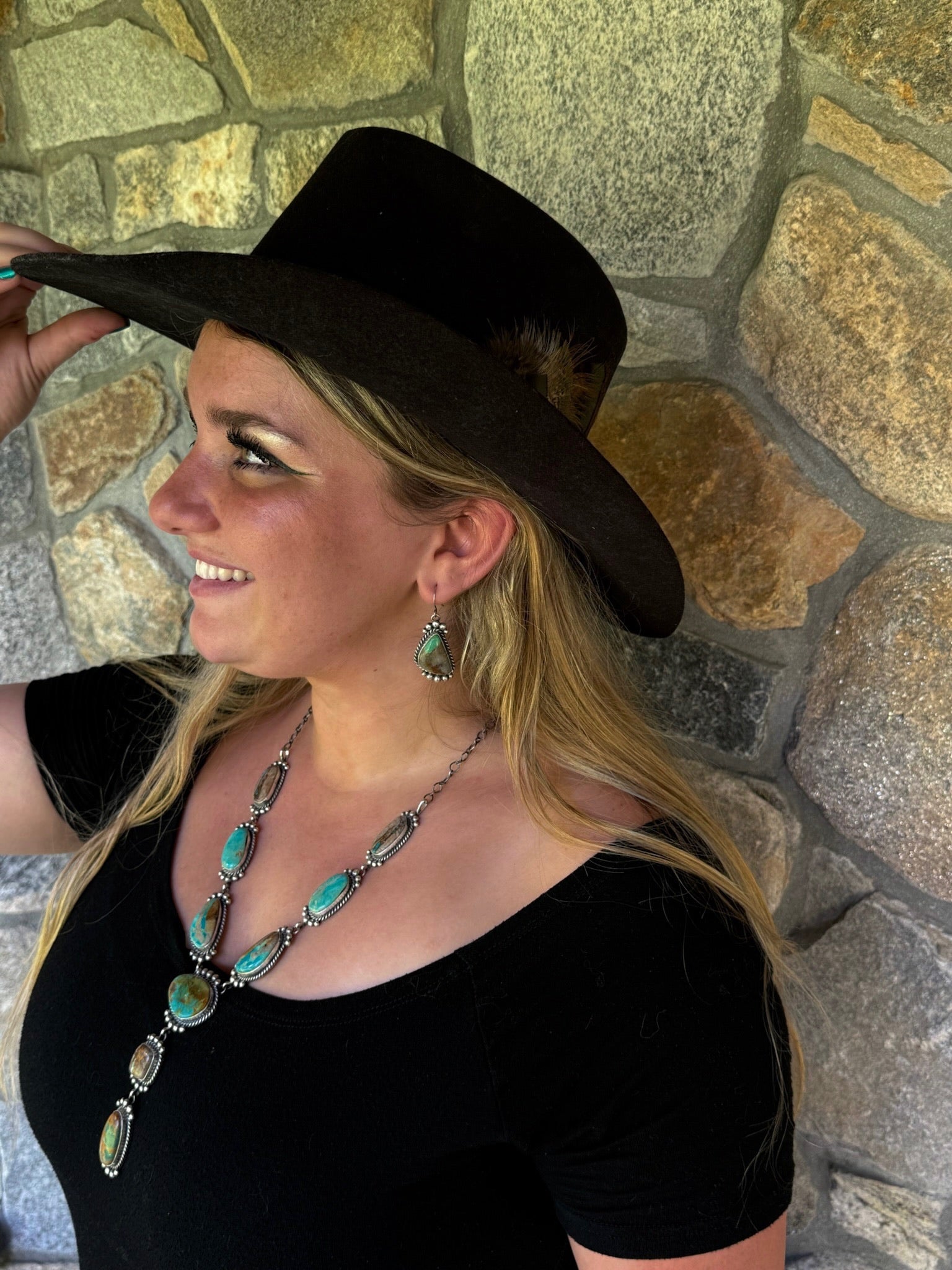 A woman wearing a black wide-brimmed cowboy hat, a black velvet top, and a turquoise and silver statement necklace, posing in front of a rustic stone wall.