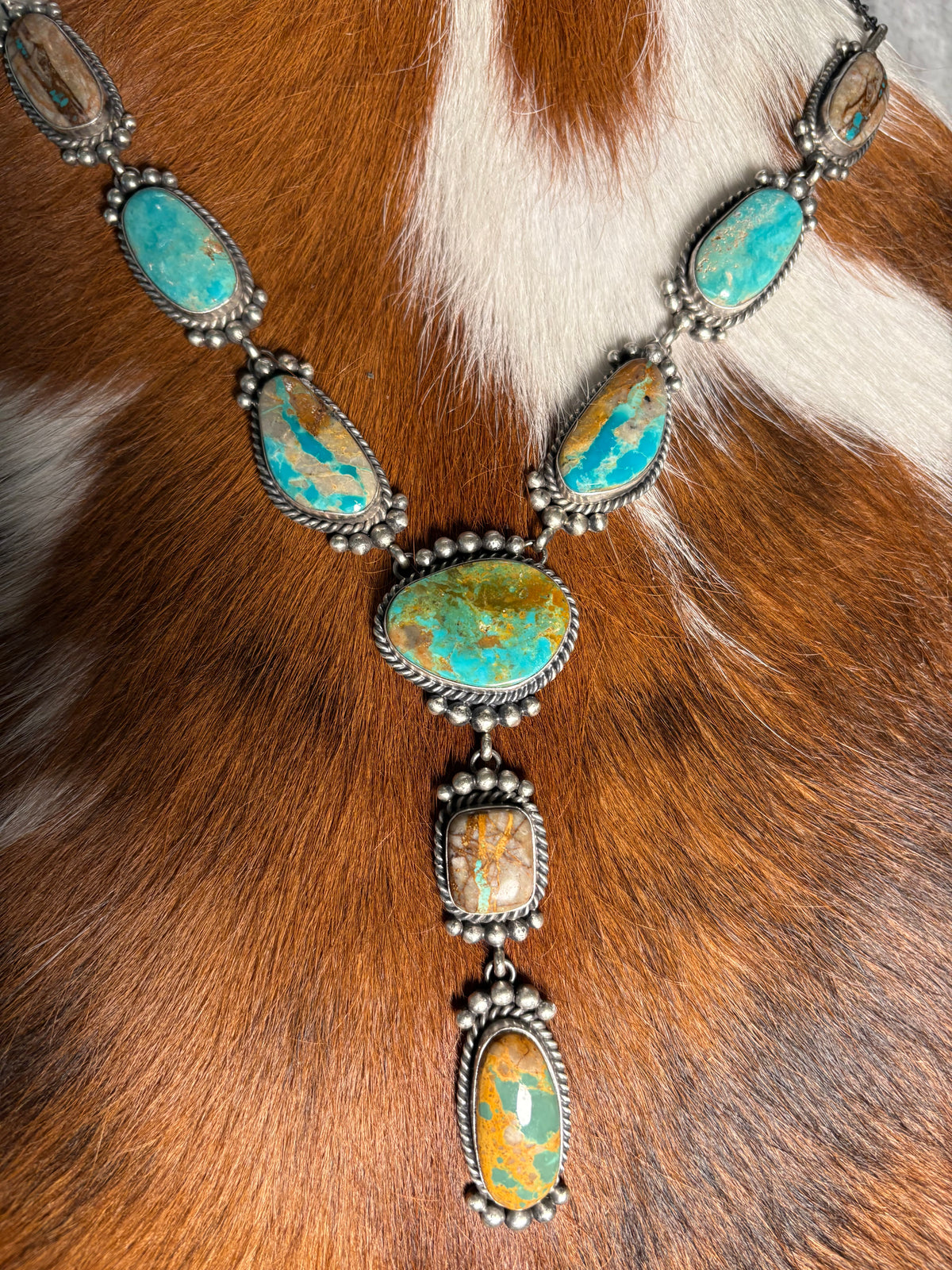 A silver-toned Southwestern-style necklace featuring multiple turquoise and orange opal stones, laid flat on a brown and white cowhide background.