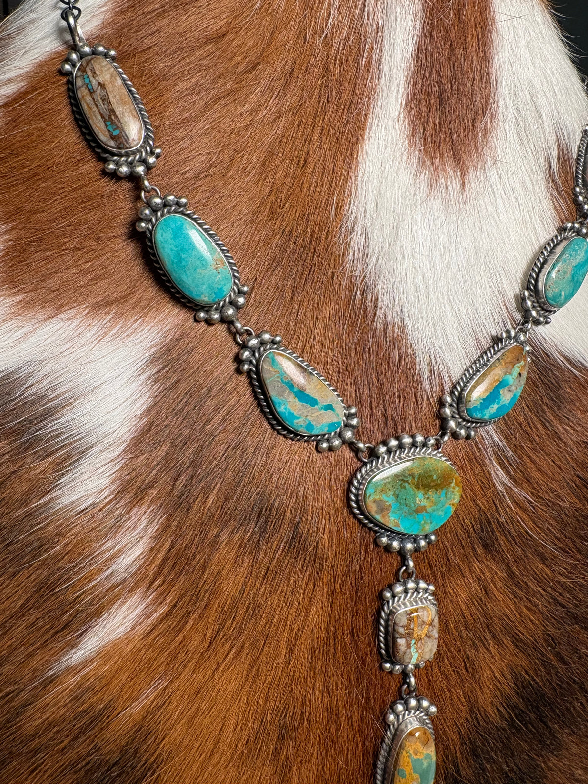A close-up of a silver-toned necklace with oval turquoise-blue stones and brown inclusions, displayed against a brown-and-white animal hide background.