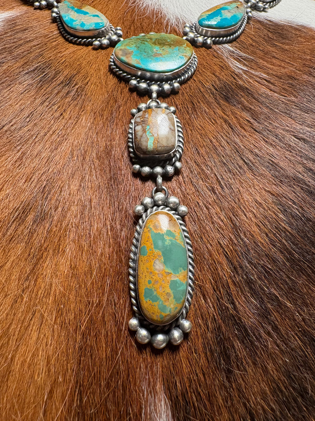 A close-up of a silver-toned necklace with multiple turquoise and orange-brown stone pendants, laid over a brown fur surface.