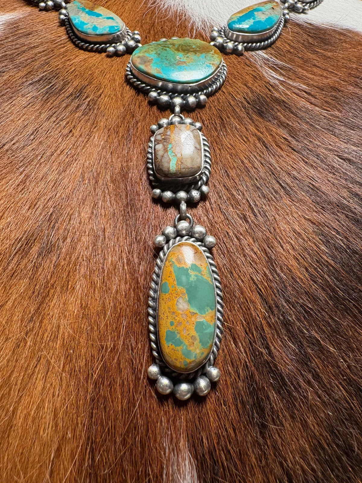 A close-up of a silver-toned necklace with multiple turquoise and orange-brown stone pendants, laid over a brown fur surface.