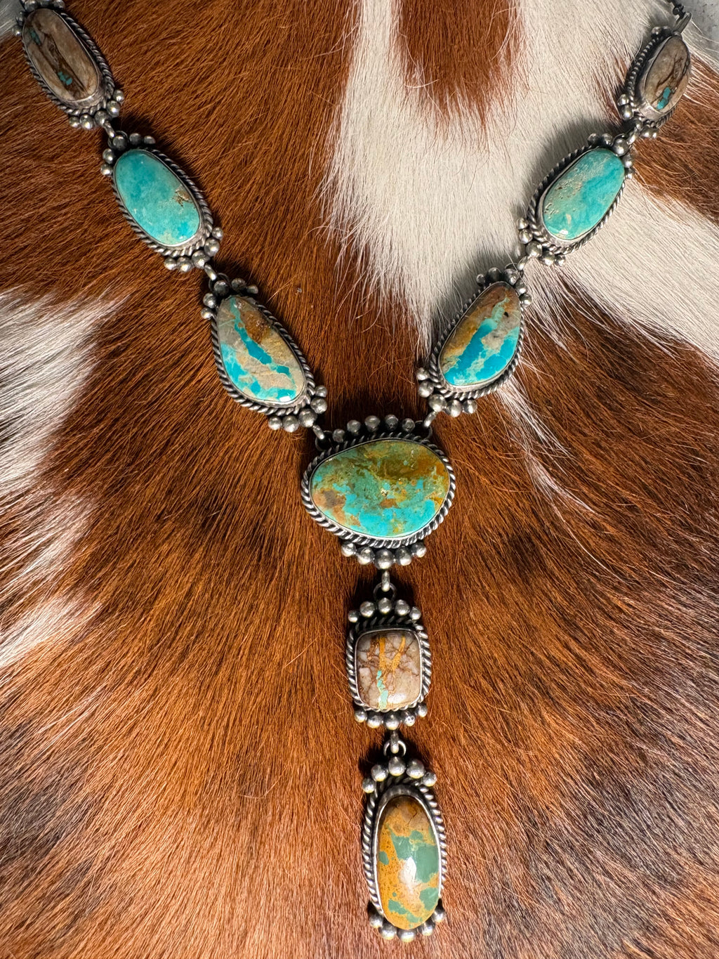 A silver-toned Southwestern-style necklace featuring multiple oval turquoise and gold marbled stones, displayed on a brown and white cowhide background.