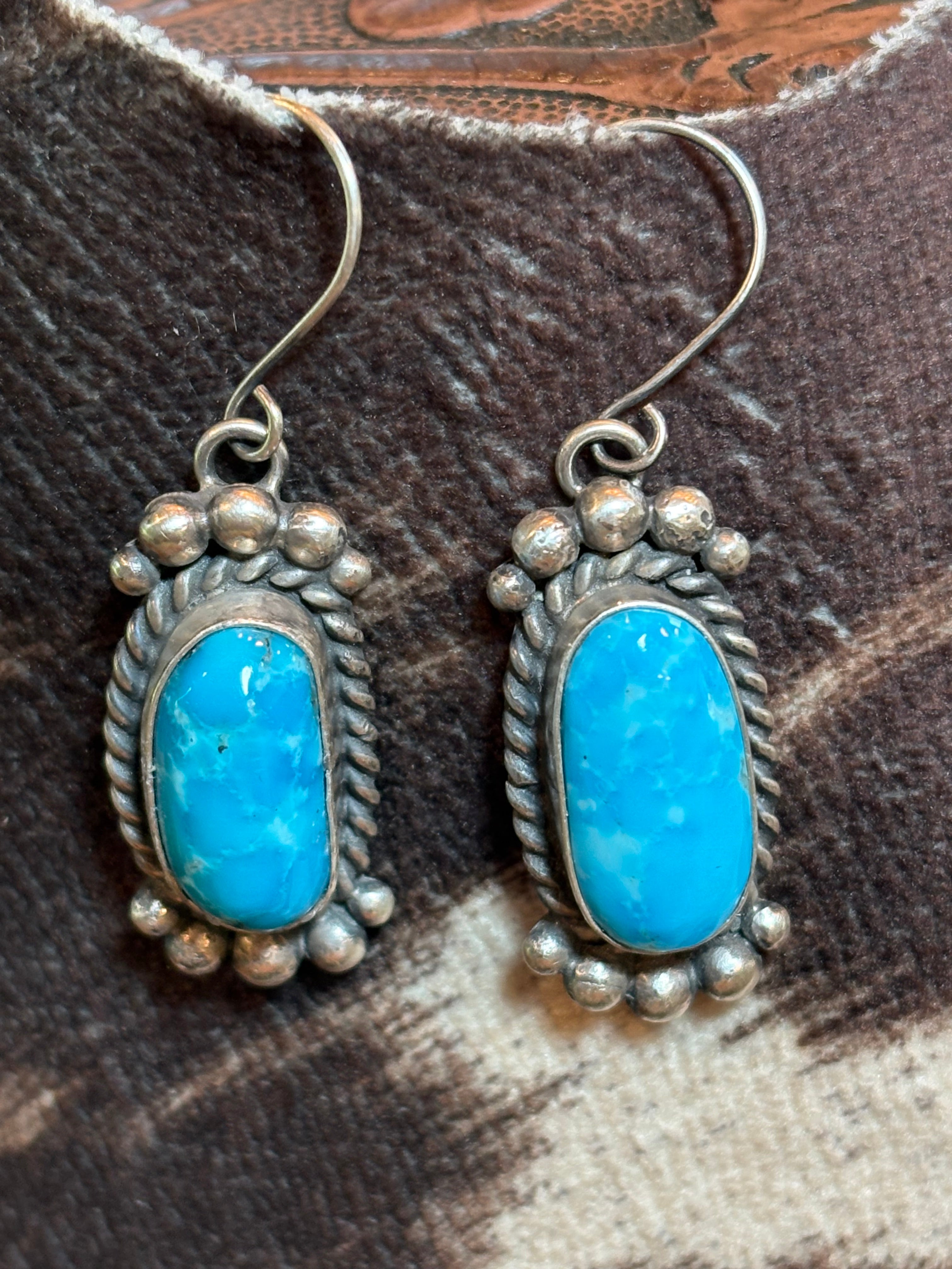 A pair of silver dangle earrings with oval turquoise stones, displayed on a textured brown and beige leather surface.