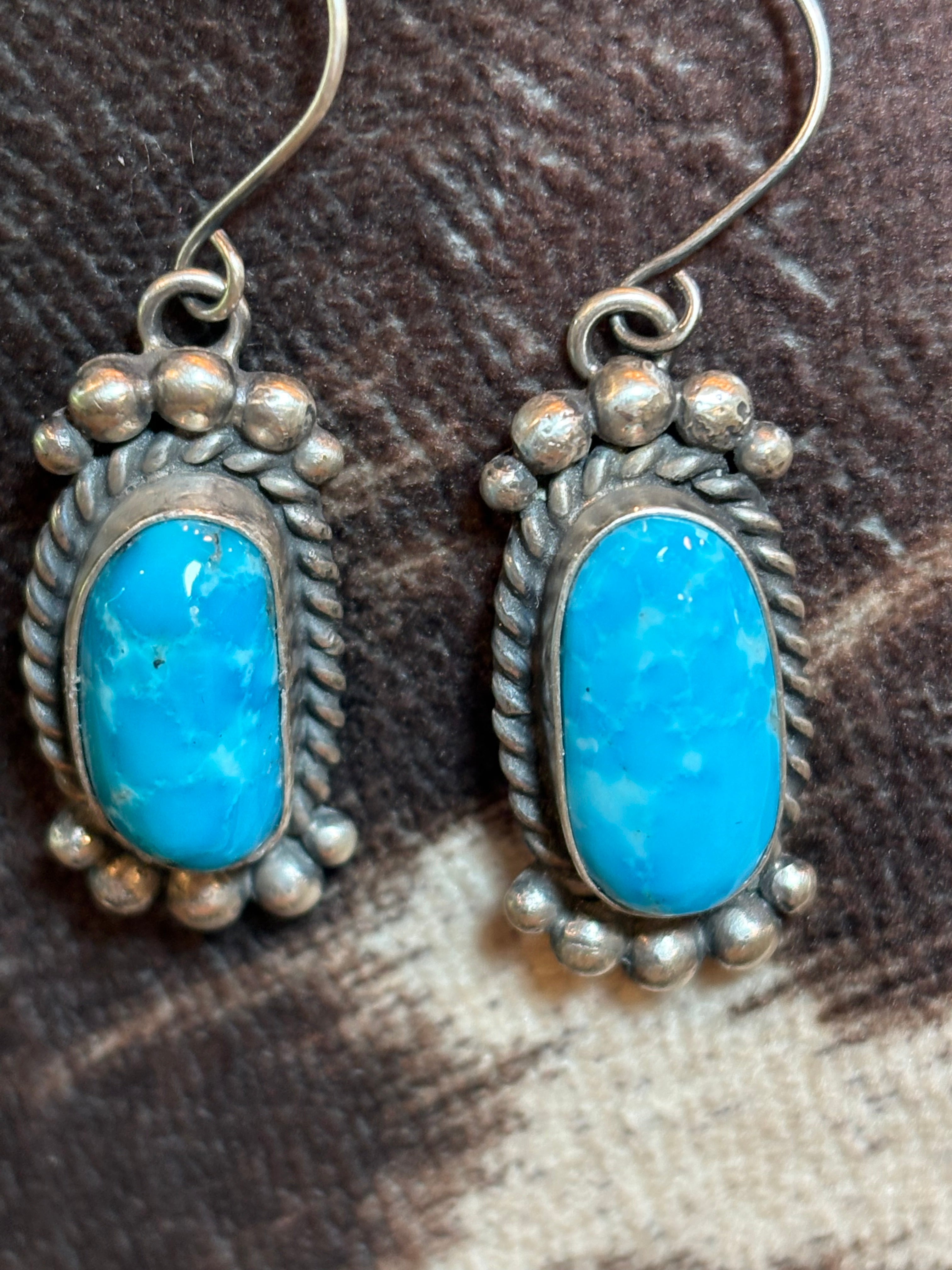 A pair of silver dangle earrings with oval turquoise stones, resting on a brown and beige textured surface.