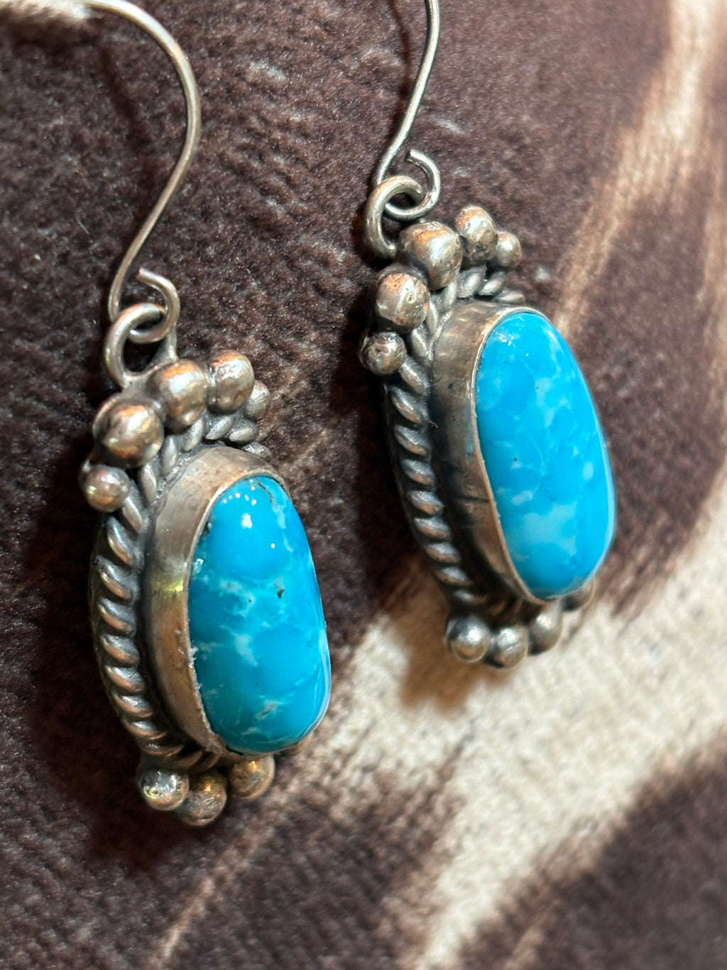 A pair of silver-toned drop earrings featuring oval turquoise blue stones, displayed on a brown and beige patterned fabric background.