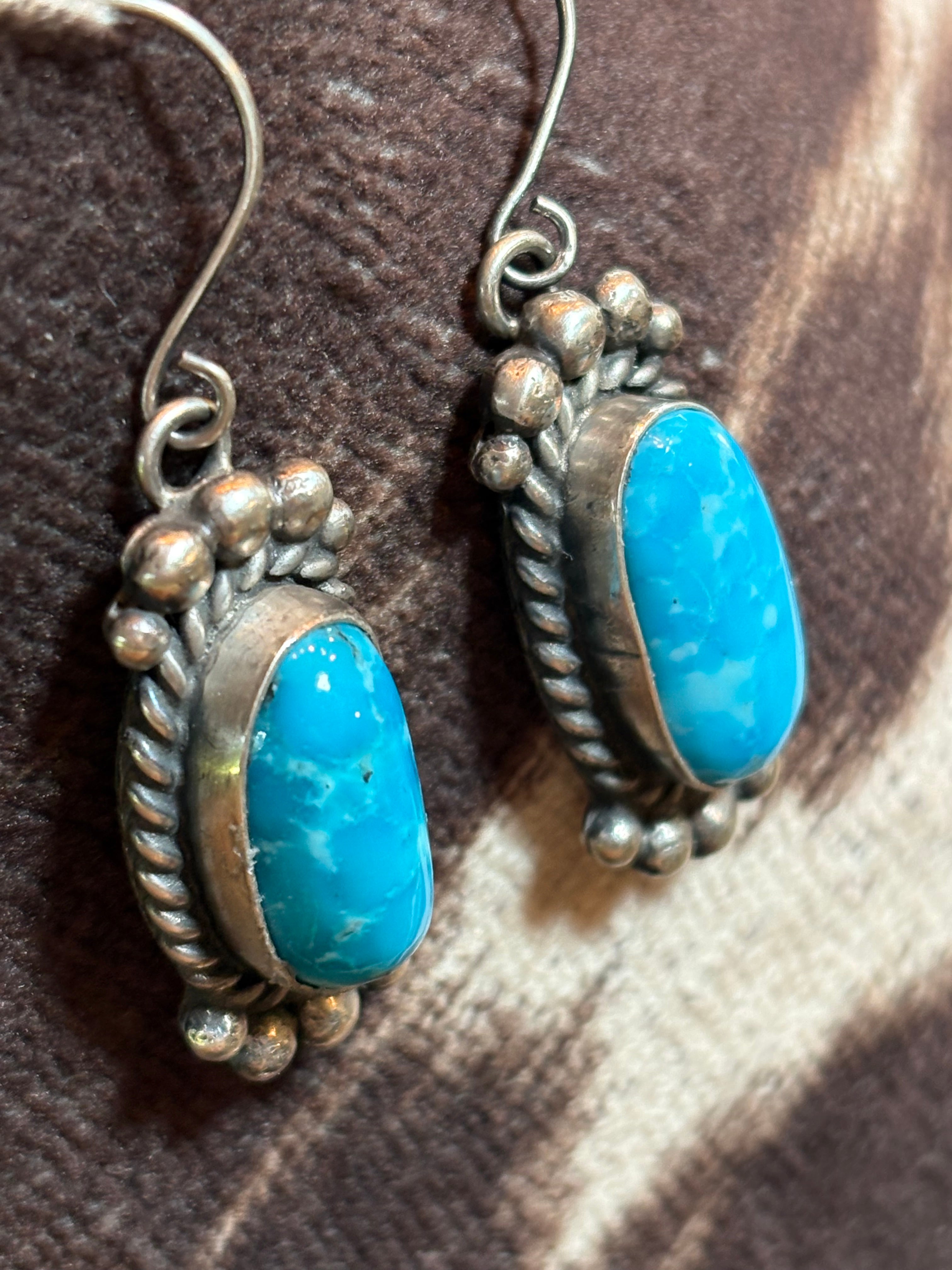 A pair of silver-toned drop earrings featuring oval turquoise blue stones, displayed on a brown and beige patterned fabric background.