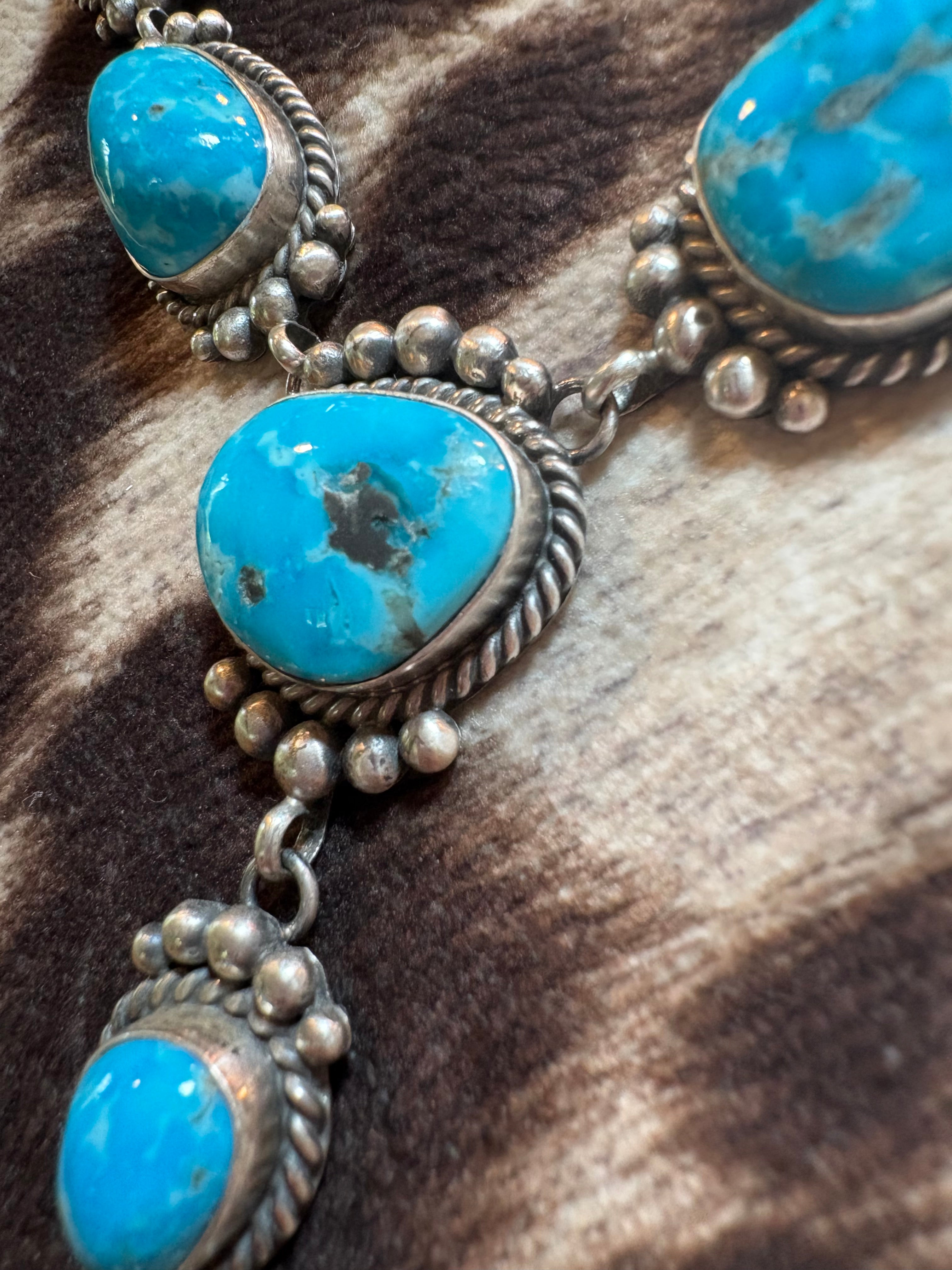 A close-up of a silver-toned jewelry piece featuring three large oval turquoise stones with dark matrix, set in textured silver bezels with small silver beads, resting on a brown and black zebra-patterned surface.
