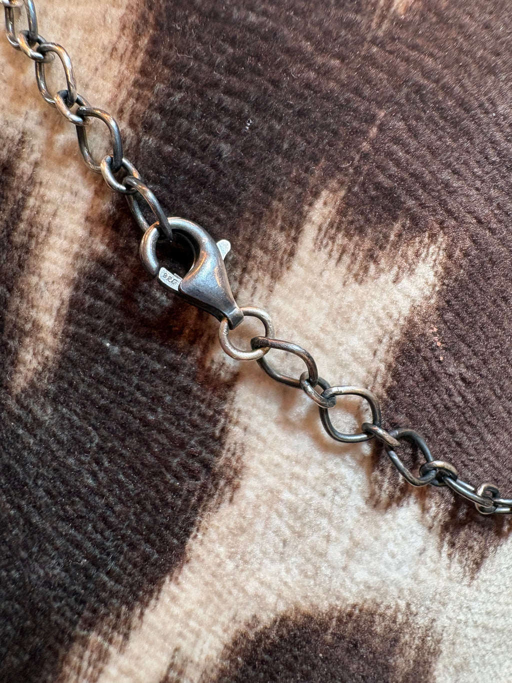 A close-up of a dark gunmetal chain with a lobster clasp, resting on a brown and beige patterned fabric background.