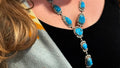 A close-up lifestyle shot of a woman wearing a silver-toned Southwestern-style necklace and matching drop earring, both featuring vibrant turquoise blue stones, paired with a black top and a patterned knit blanket.