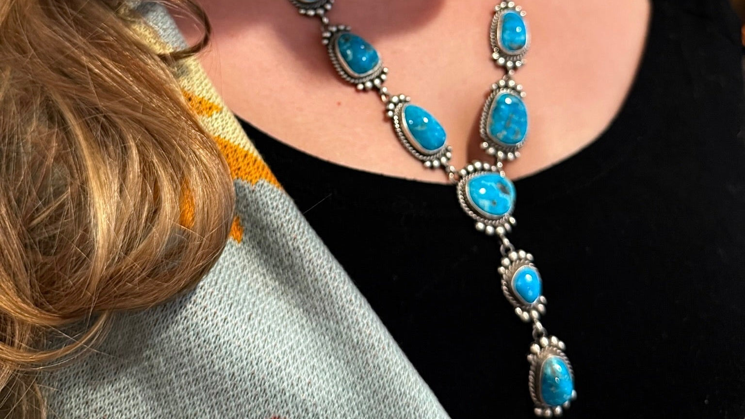 A close-up lifestyle shot of a woman wearing a silver-toned Southwestern-style necklace and matching drop earring, both featuring vibrant turquoise blue stones, paired with a black top and a patterned knit blanket.