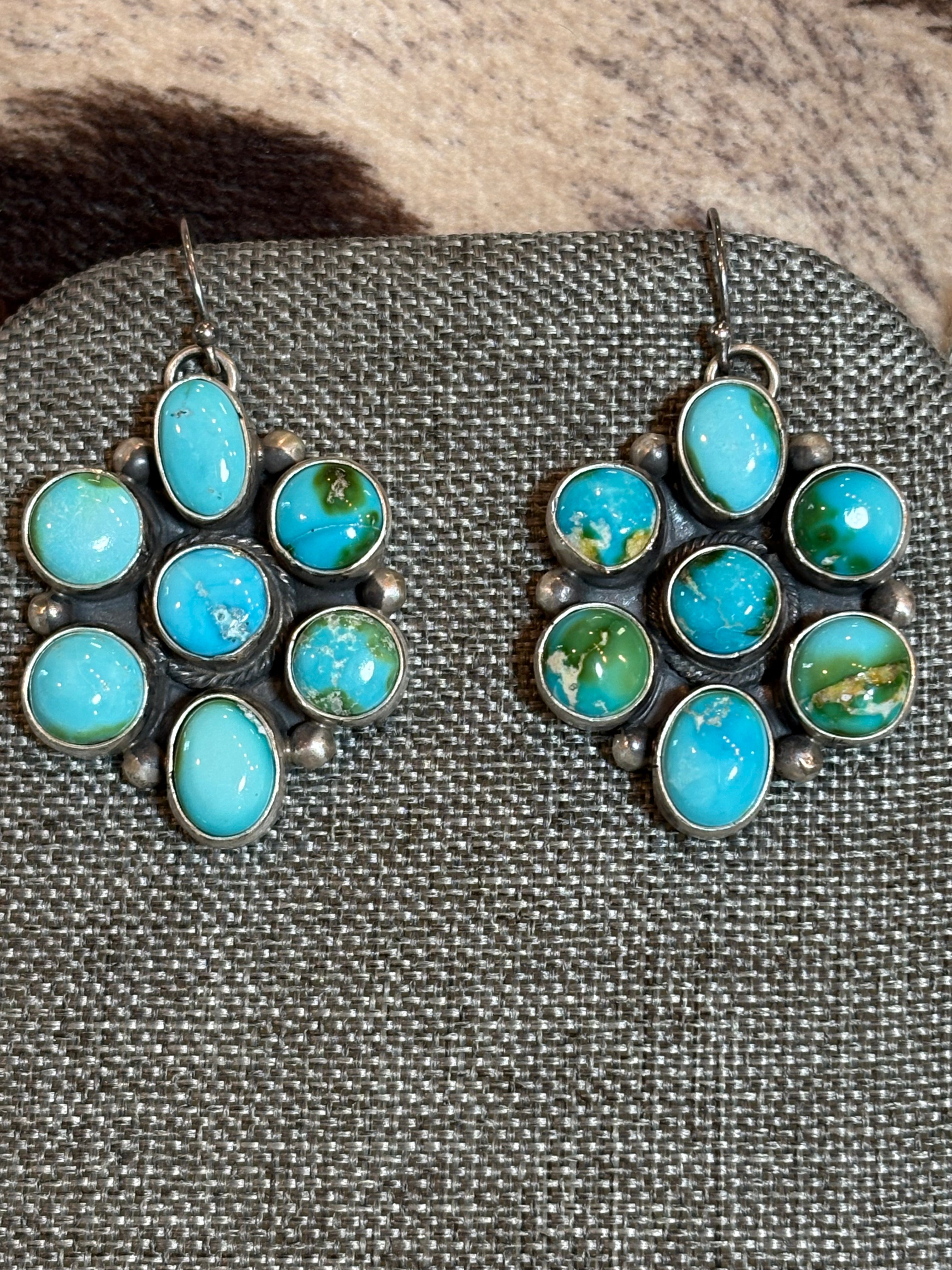 A pair of silver-toned dangle earrings with oval turquoise-blue stones and several round green marbled turquoise accents, displayed on a textured gray fabric surface.