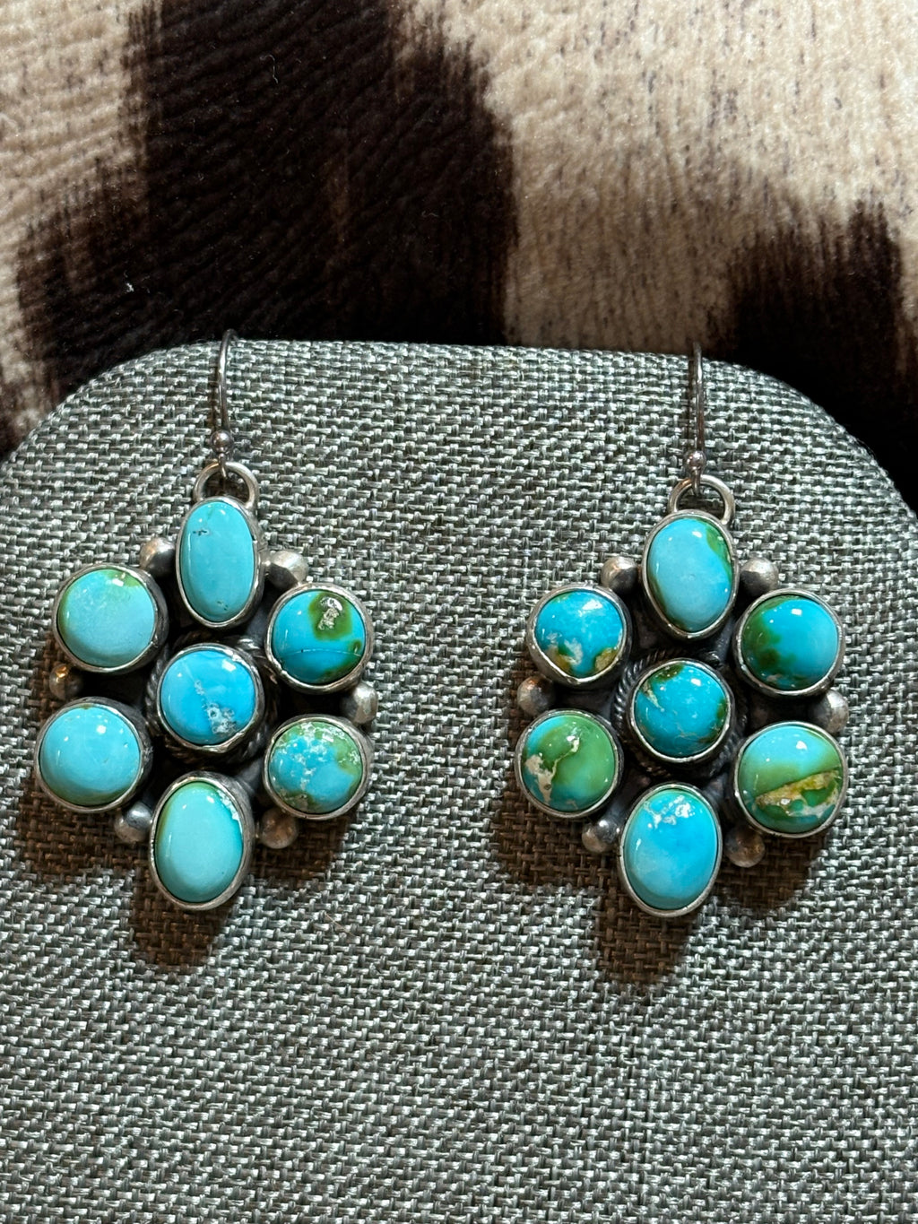A pair of silver dangle earrings with oval turquoise and green marbled stones, displayed on a textured gray jewelry stand.