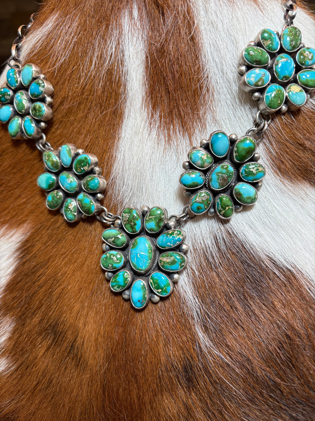A close-up of a silver-toned Southwestern-style necklace featuring clusters of turquoise stones, laid flat on a brown and white cowhide background.