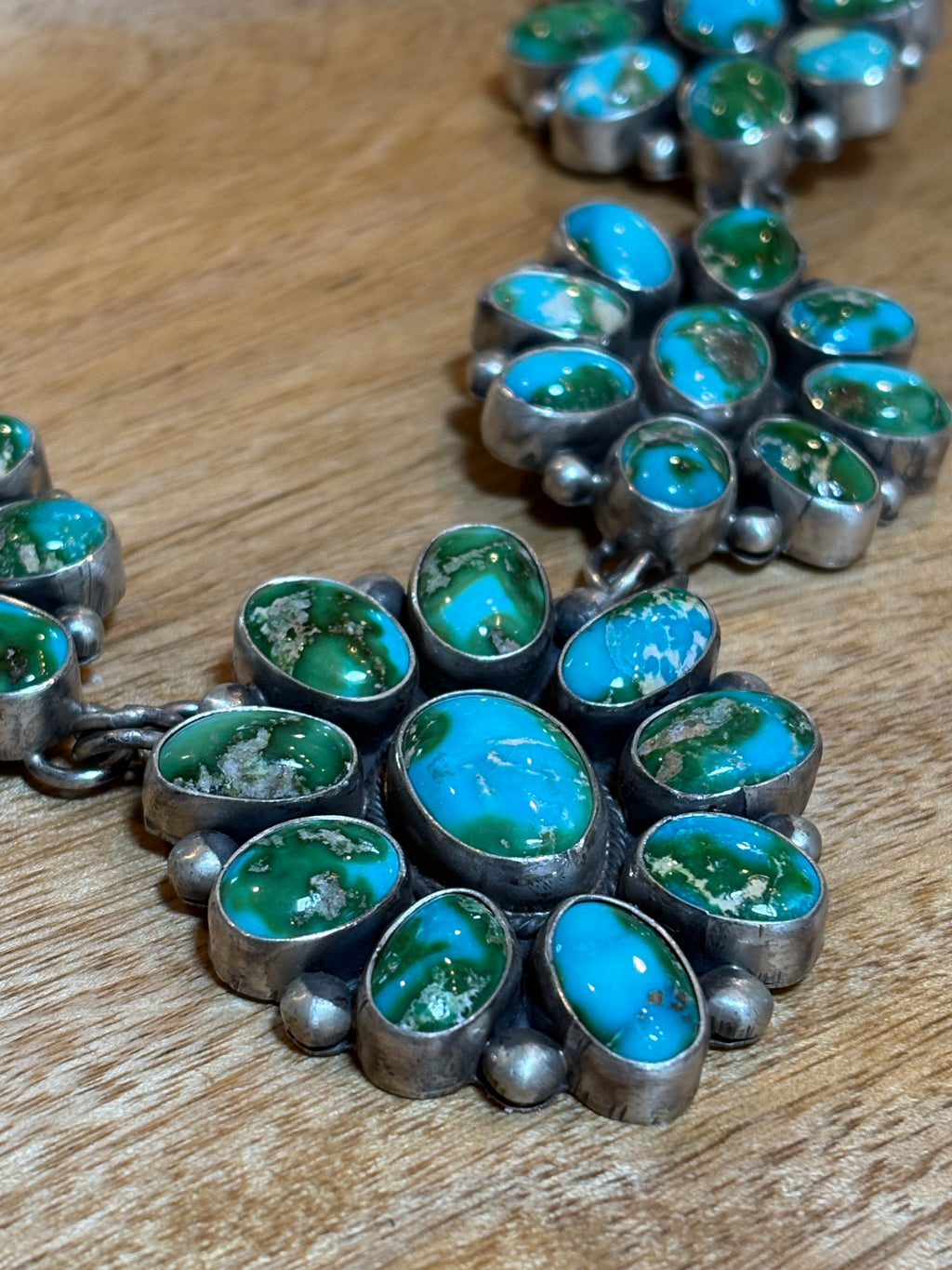 A close-up of a silver-toned jewelry piece featuring multiple oval turquoise and green stone clusters set in silver, resting on a wooden surface.