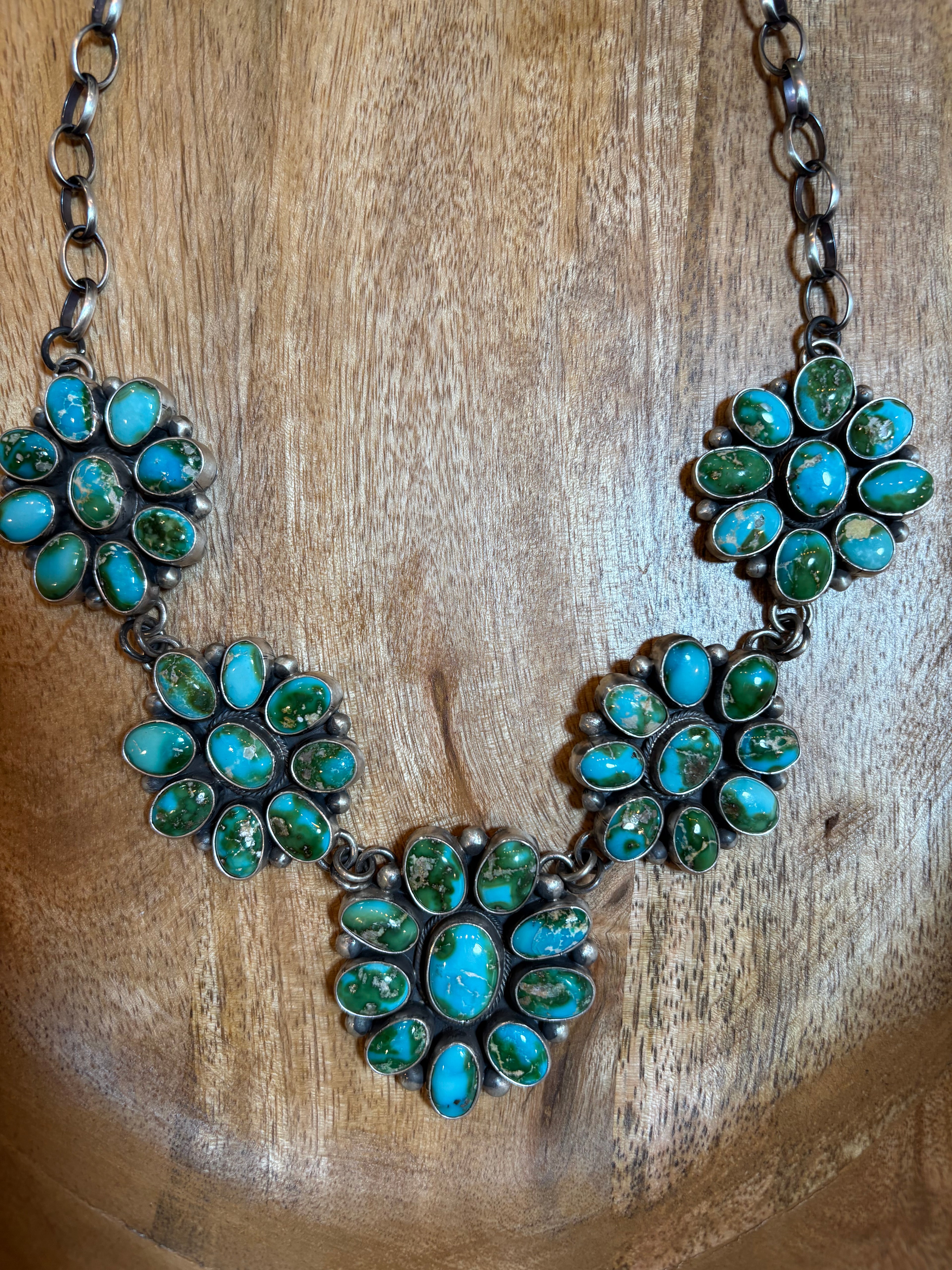 A silver-toned statement necklace featuring five floral clusters of turquoise stones in different shades, displayed on a warm wooden surface.