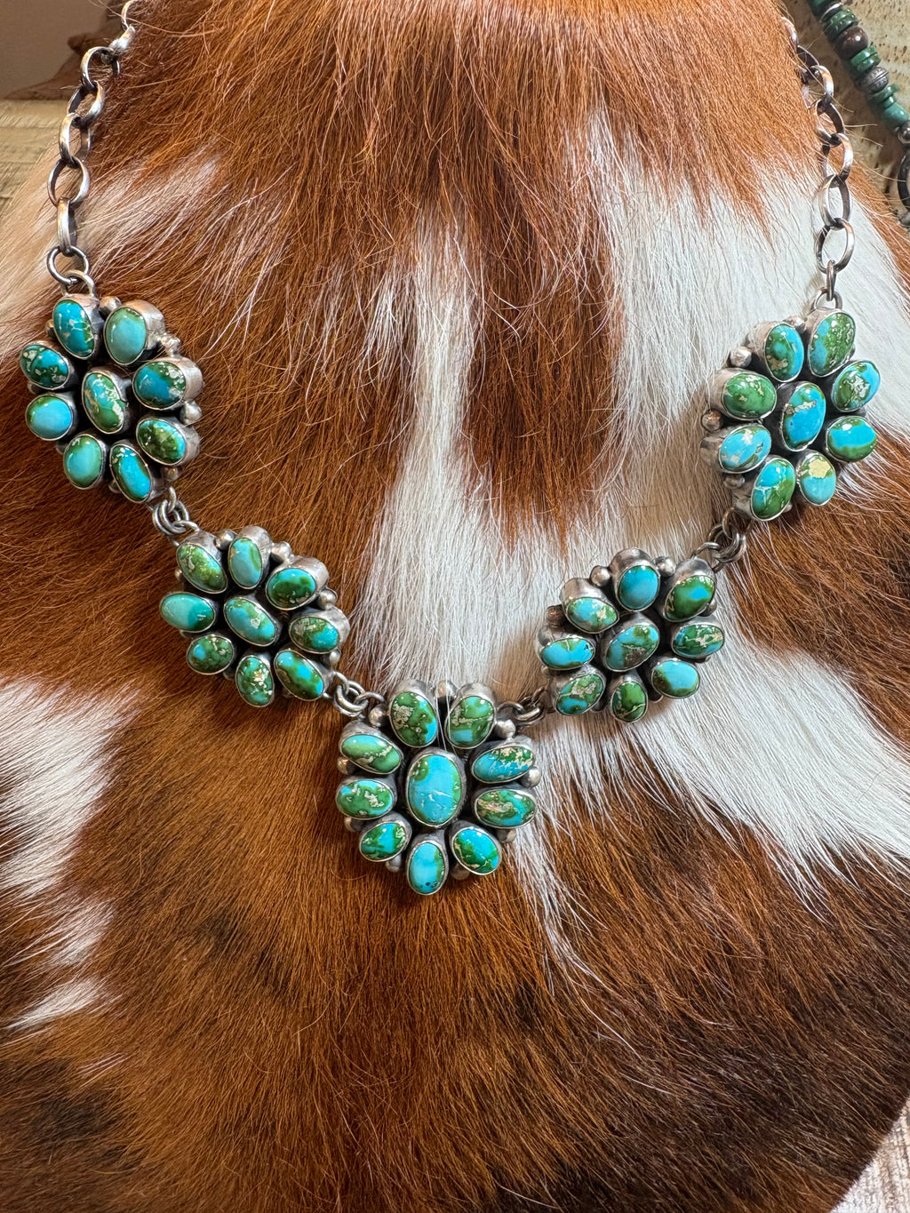 A silver-toned statement necklace featuring multiple clusters of turquoise stones, displayed on a brown and white cowhide background.