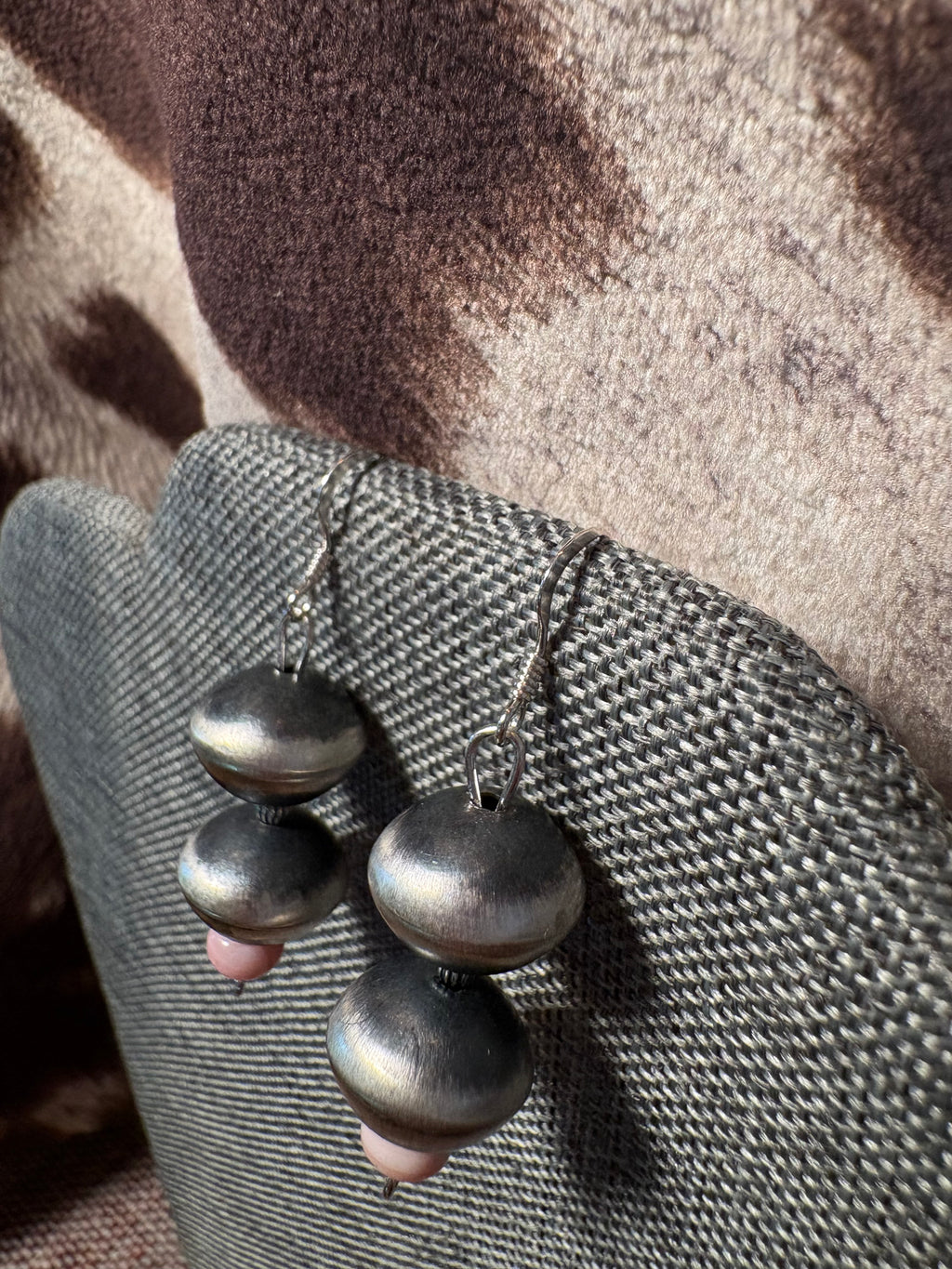A pair of handmade dangle earrings with stacked metallic silver beads and small pink stone accents, displayed on a textured gray fabric stand.