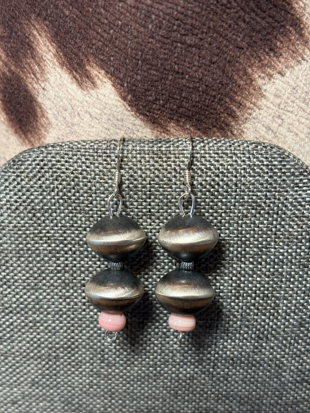 A pair of handmade dangle earrings with dark oxidized metal beads and small pink spacer beads, displayed on a textured gray fabric surface.