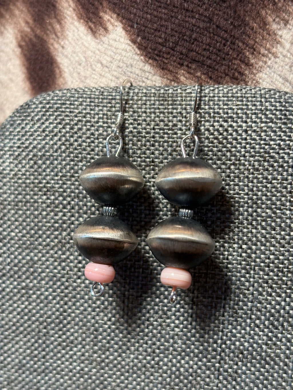 A pair of handmade dangle earrings featuring dark metallic bronze beads and small pink spacer beads, displayed on a textured gray fabric surface.