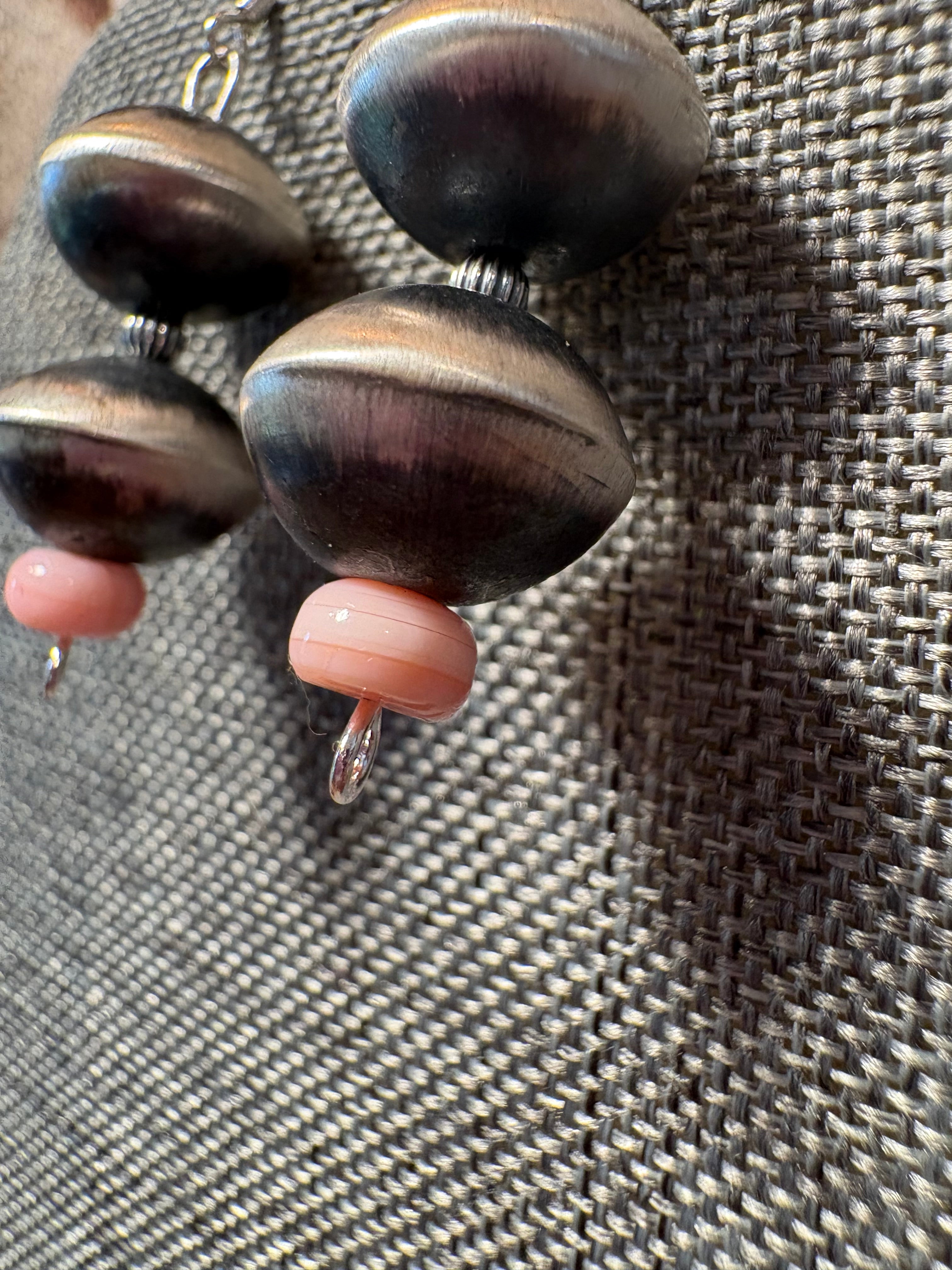 Close-up of handmade beaded earrings with dark brown and black wood beads accented by light pink coral beads, displayed on a gray woven fabric background.