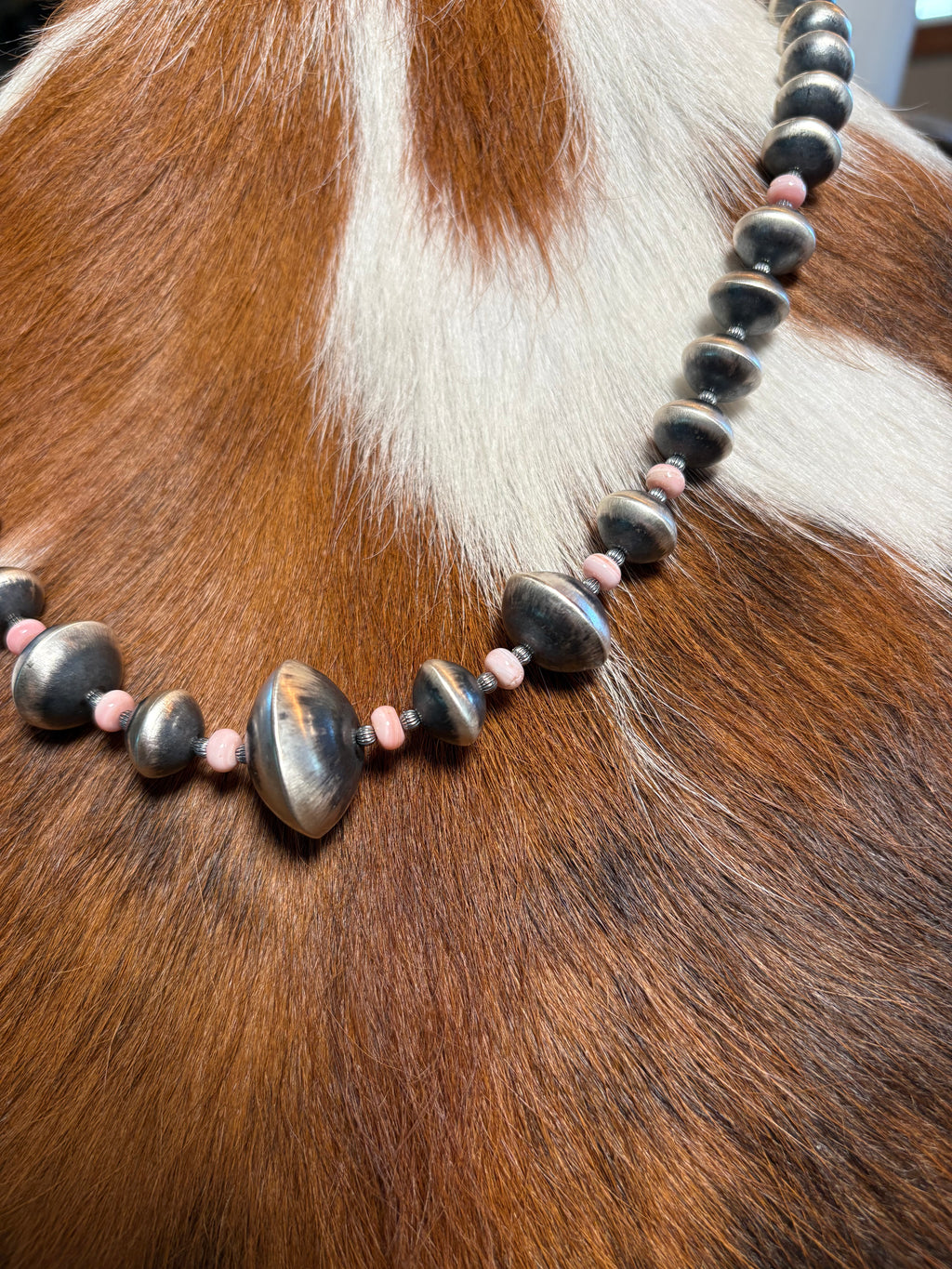 A close-up of a beaded necklace draped over brown and white cowhide. The necklace features large polished dark gray stone beads with smaller pink spacer beads.