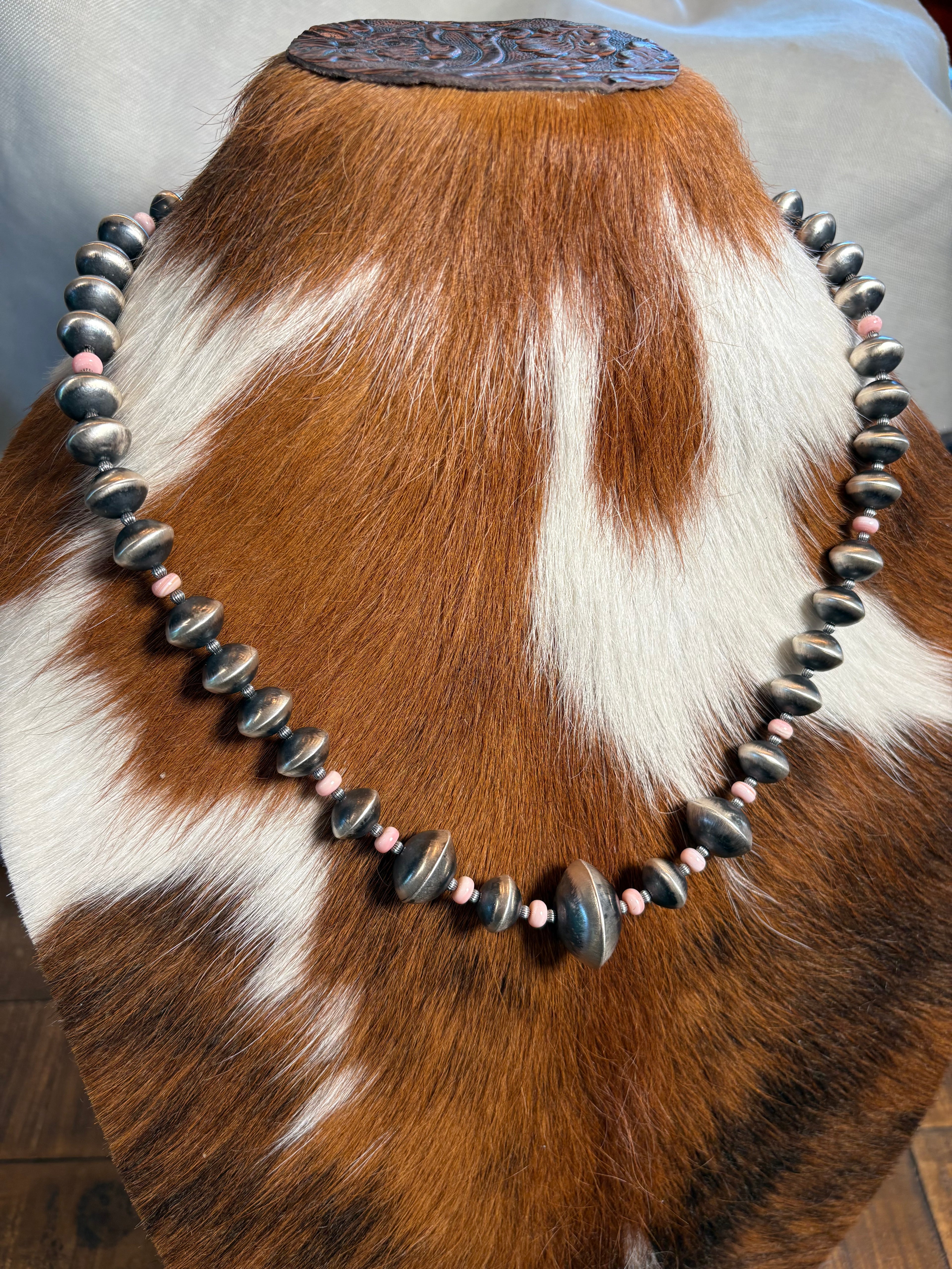 A beaded necklace displayed on a cowhide mannequin. The necklace features large polished dark gray stone beads and smaller pink accent beads.
