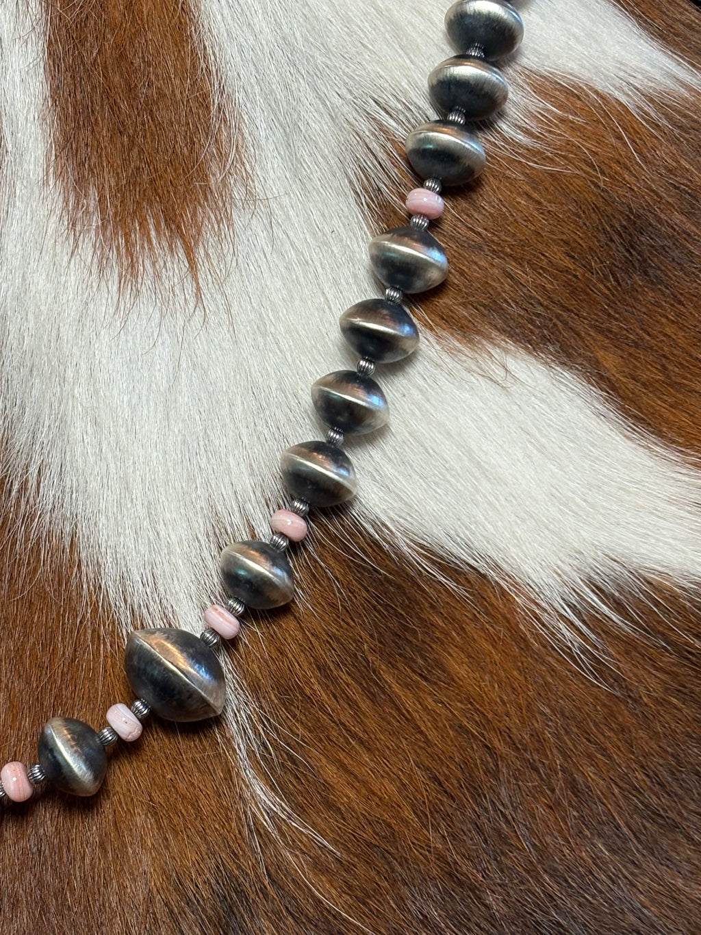 A beaded necklace featuring large, glossy dark gray beads and small pink spacer beads, laid out diagonally on a brown and white cowhide background.