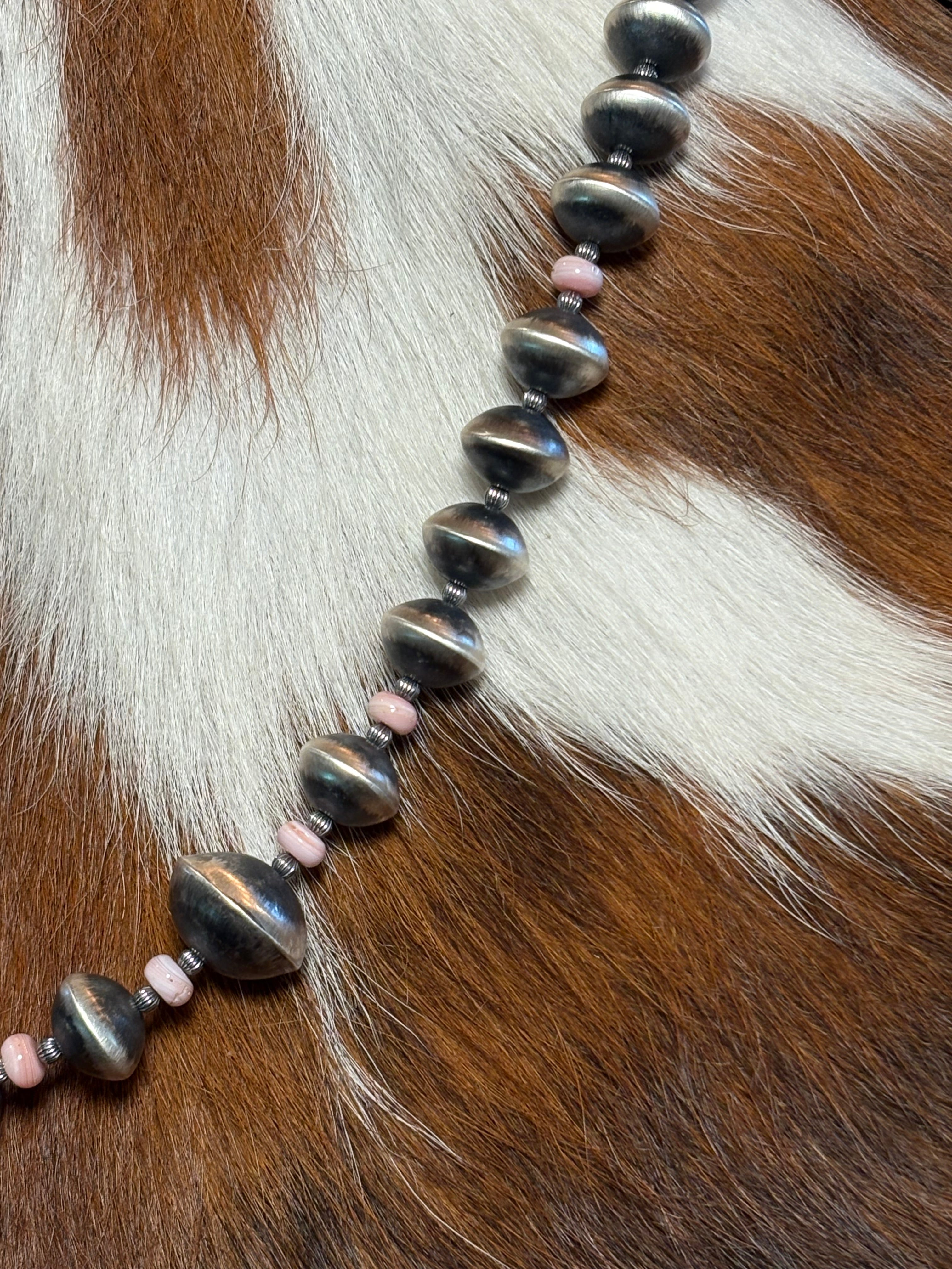 A beaded necklace featuring large, glossy dark gray beads and small pink spacer beads, laid out diagonally on a brown and white cowhide background.
